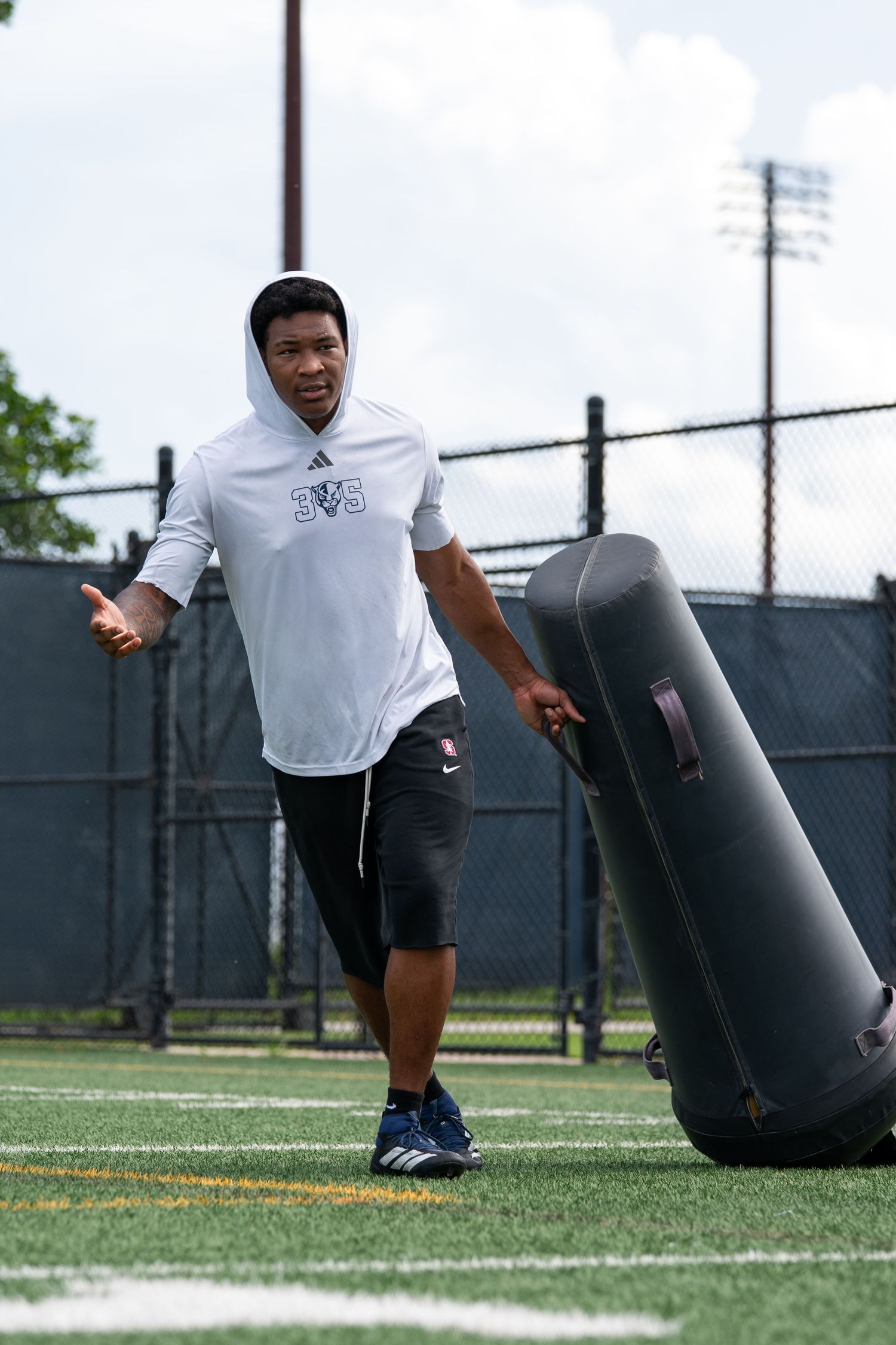 Athlete in white Adidas hoodie pushes blocking dummy on turf football field during training drill.