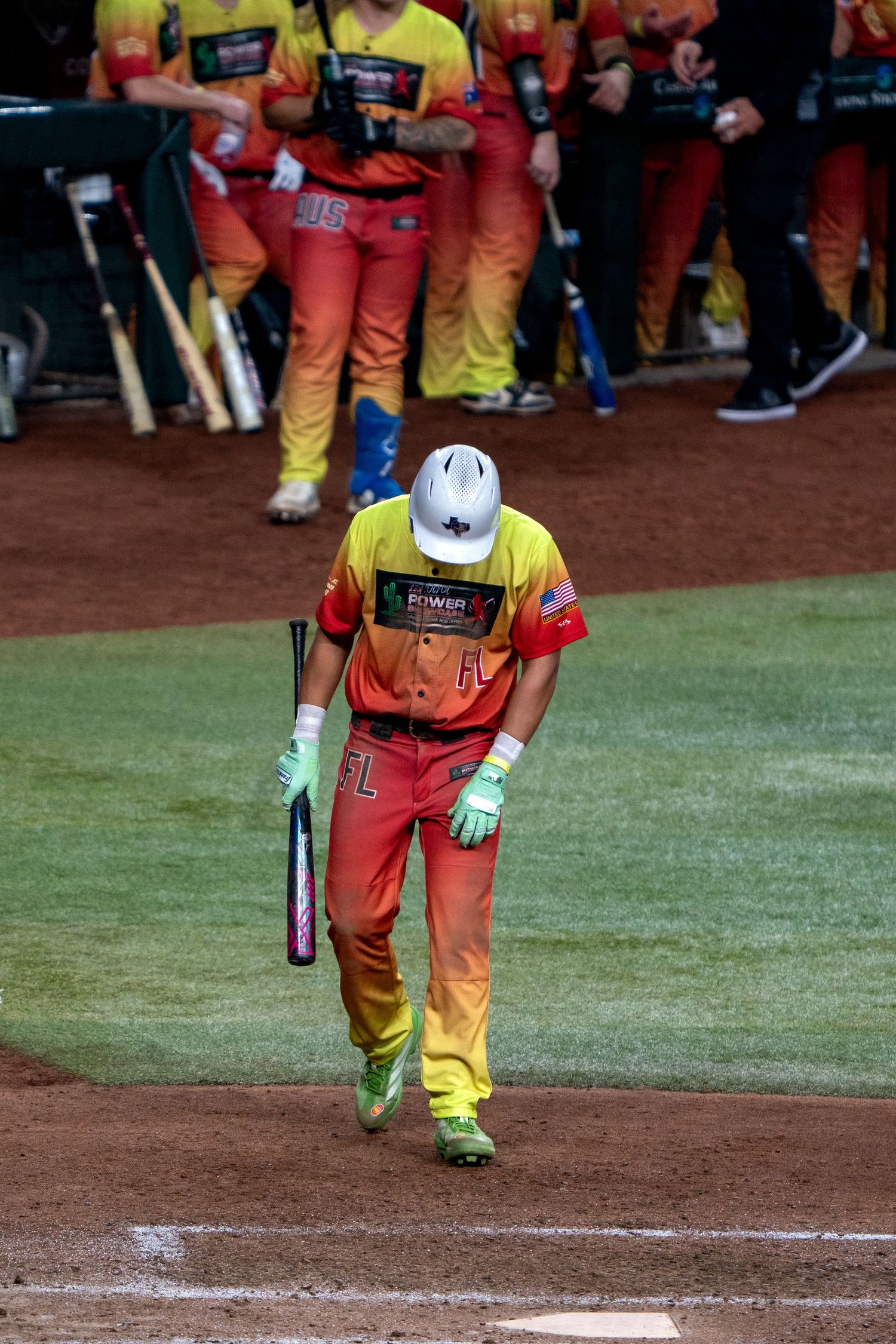 Baseball player in orange and yellow uniform bends down to place a bat on home plate during a game.