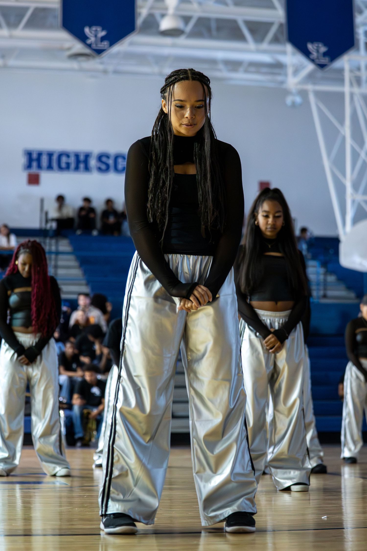 Teen dancers in silver pants and black tops perform in a high school gymnasium during a dance competition.