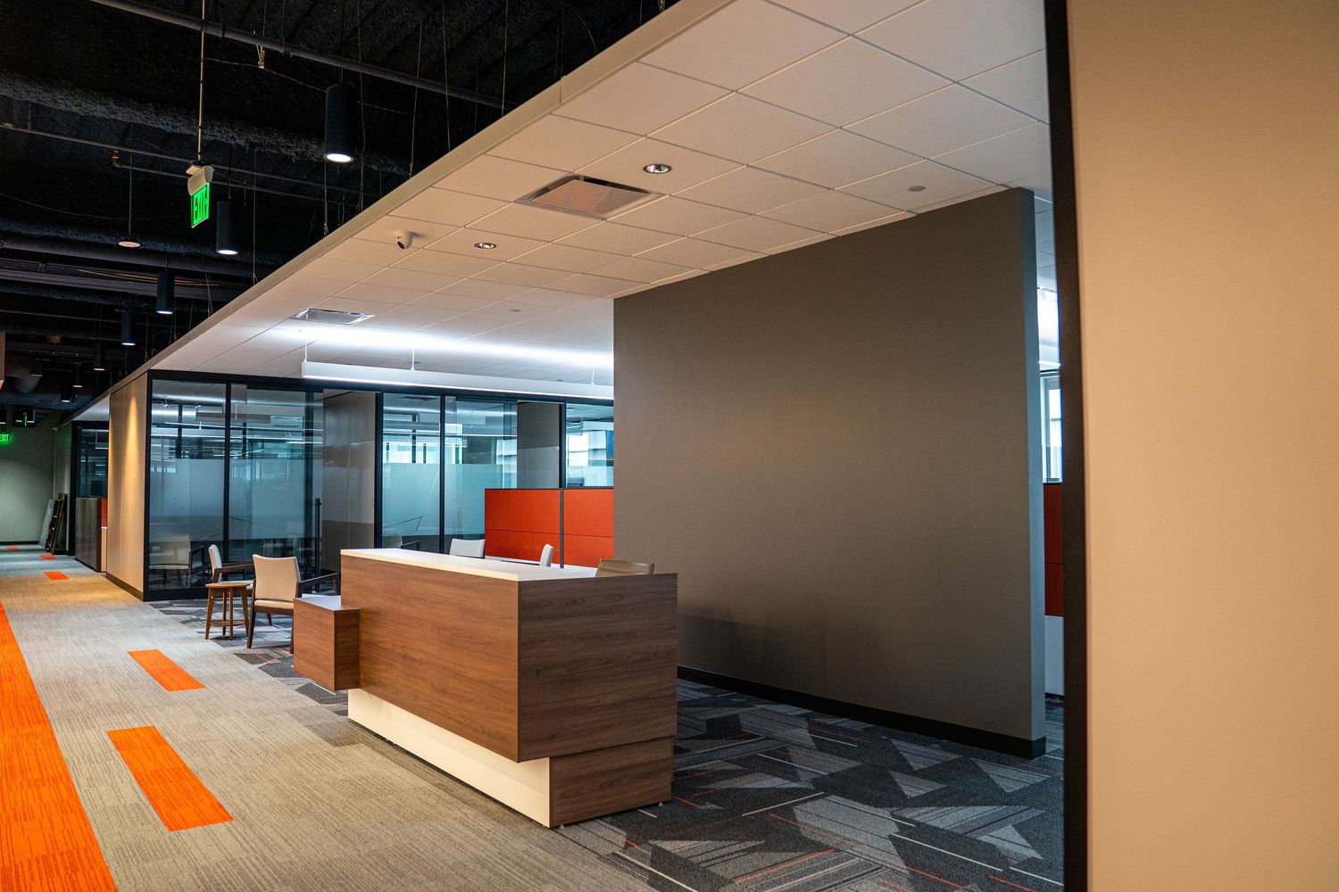 Modern office reception area with wooden desk and orange accent walls against glass partitions and patterned carpet.