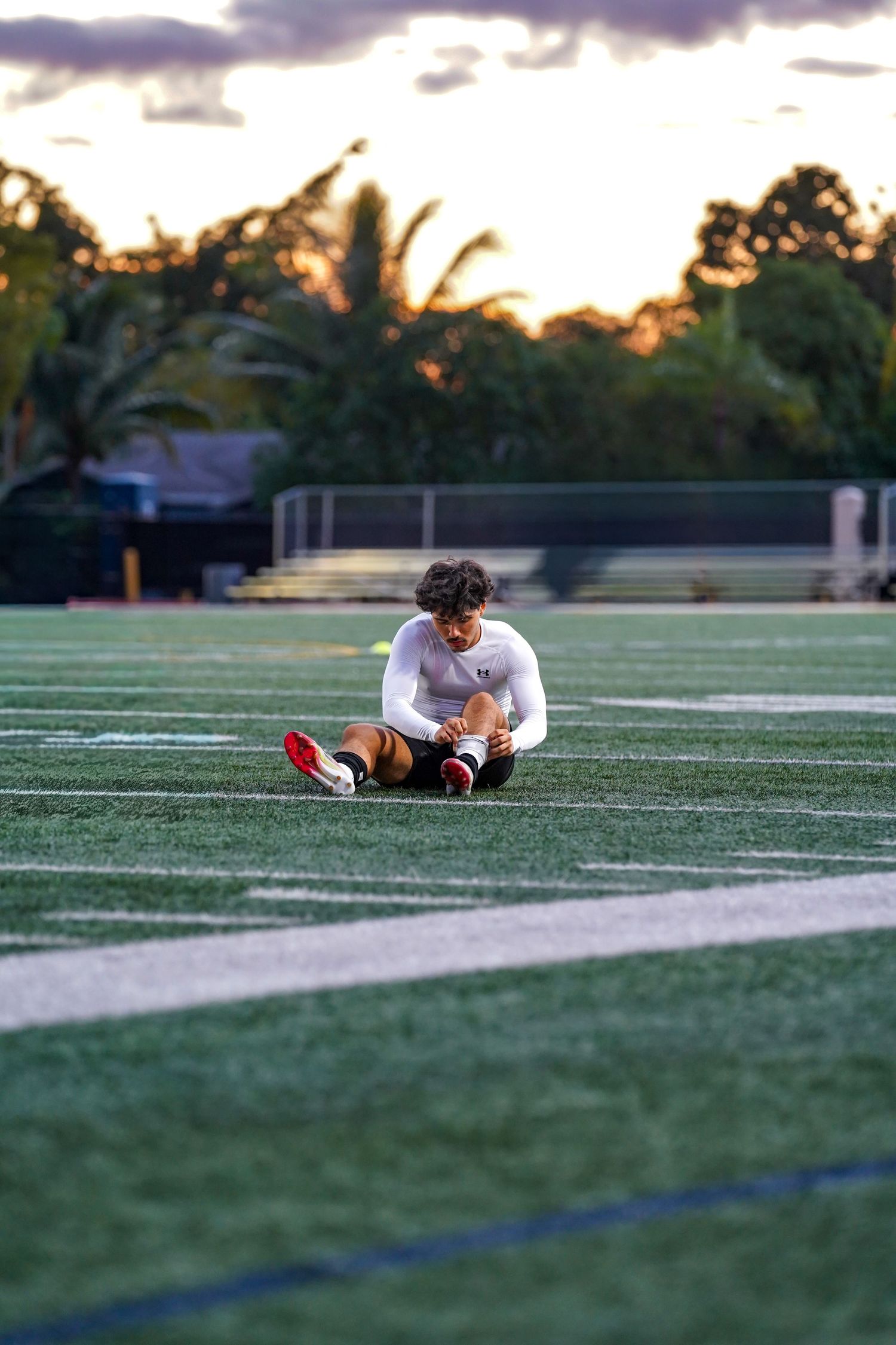 Athlete sitting alone on turf field at sunset with palm trees in background.