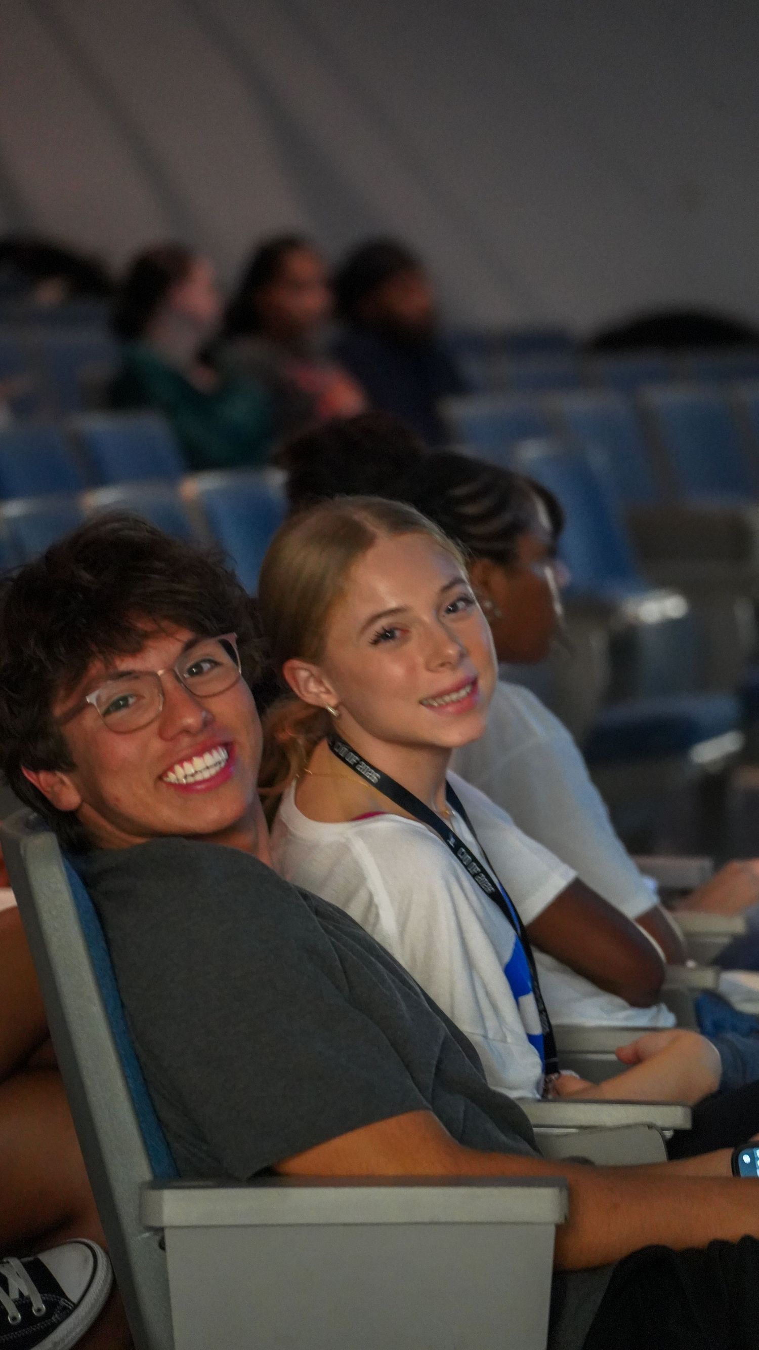 Students sit attentively in blue auditorium seats during an educational event.
