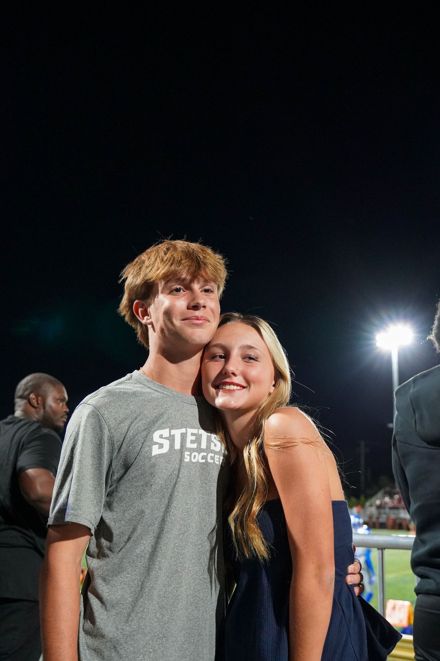 A couple sharing an intimate moment at a nighttime sporting event under stadium lights.