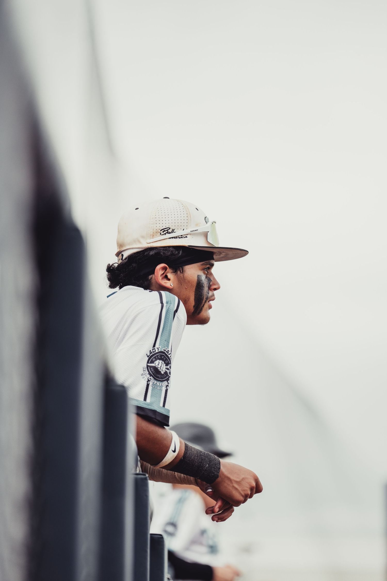 Baseball player with dreadlocks and eye black leaning on dugout railing wearing white uniform and fitted cap.