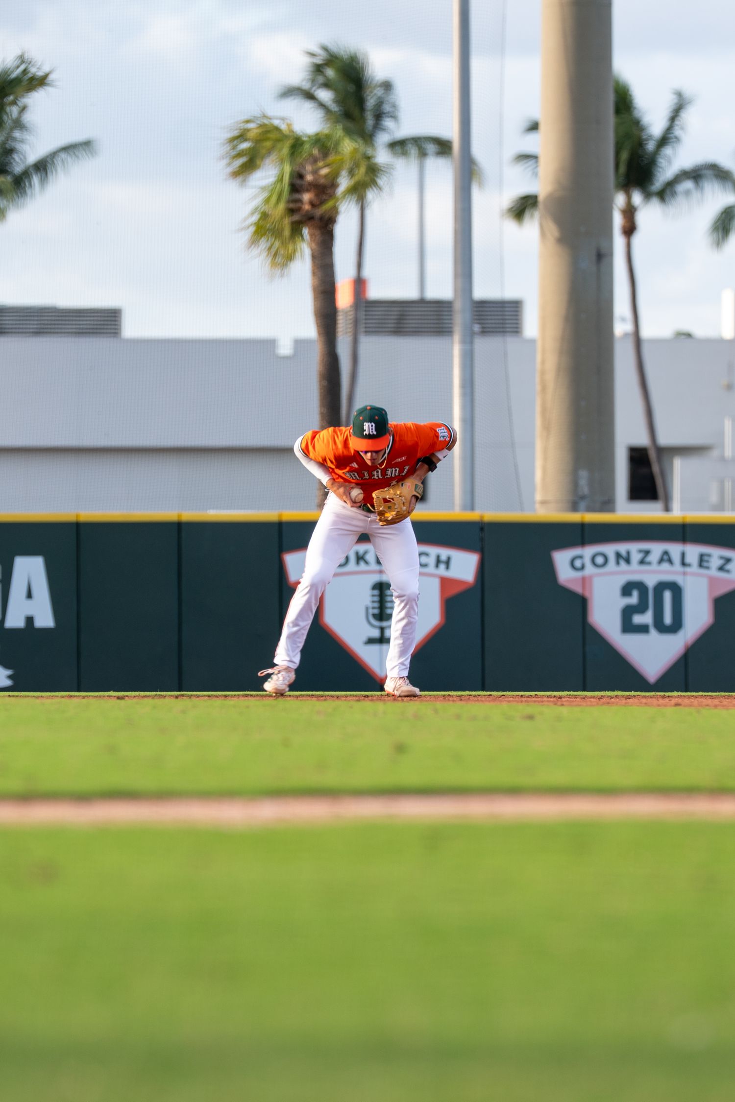 Miami Hurricanes baseball player in orange jersey fields ball near Gonzalez memorial sign with palm trees.