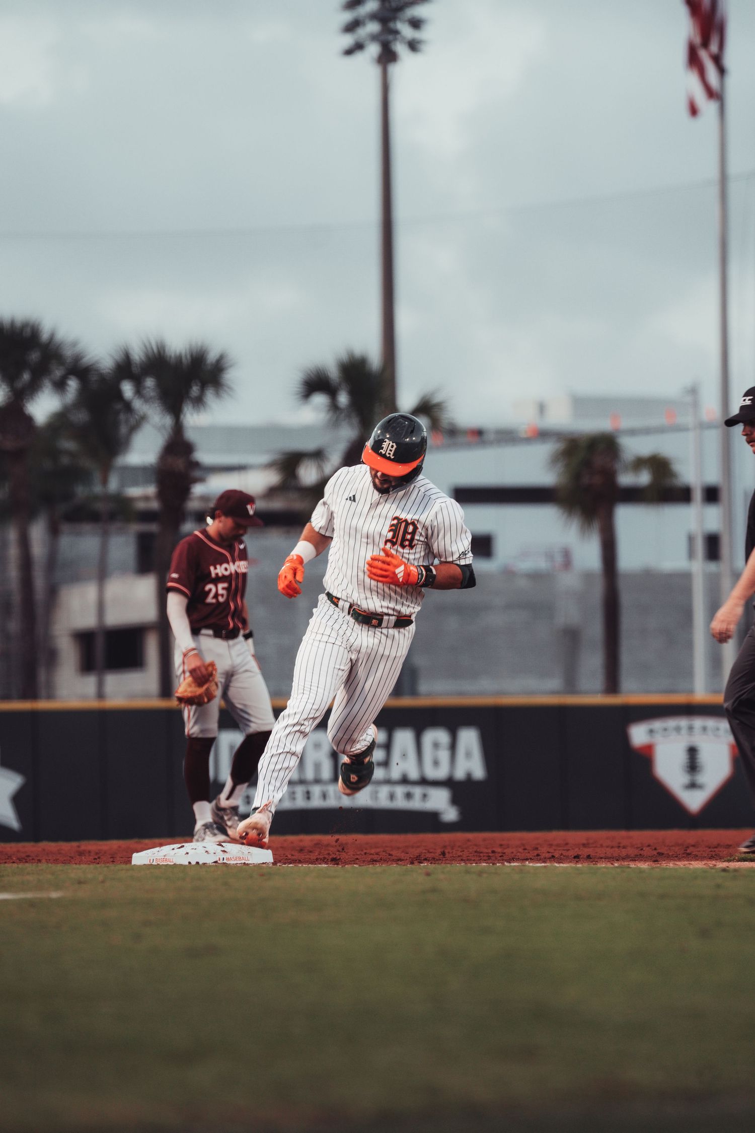 Baseball player rounds third base in white pinstripe uniform with orange gloves during a college baseball game.