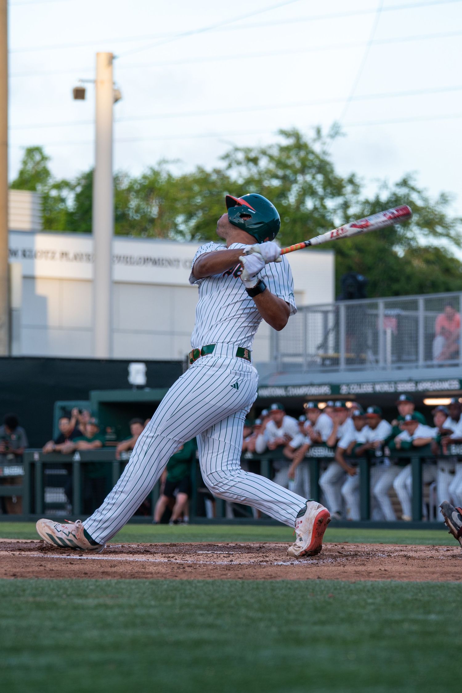 Baseball player in white pinstripe uniform swinging bat during a game, with teammates watching from the dugout.