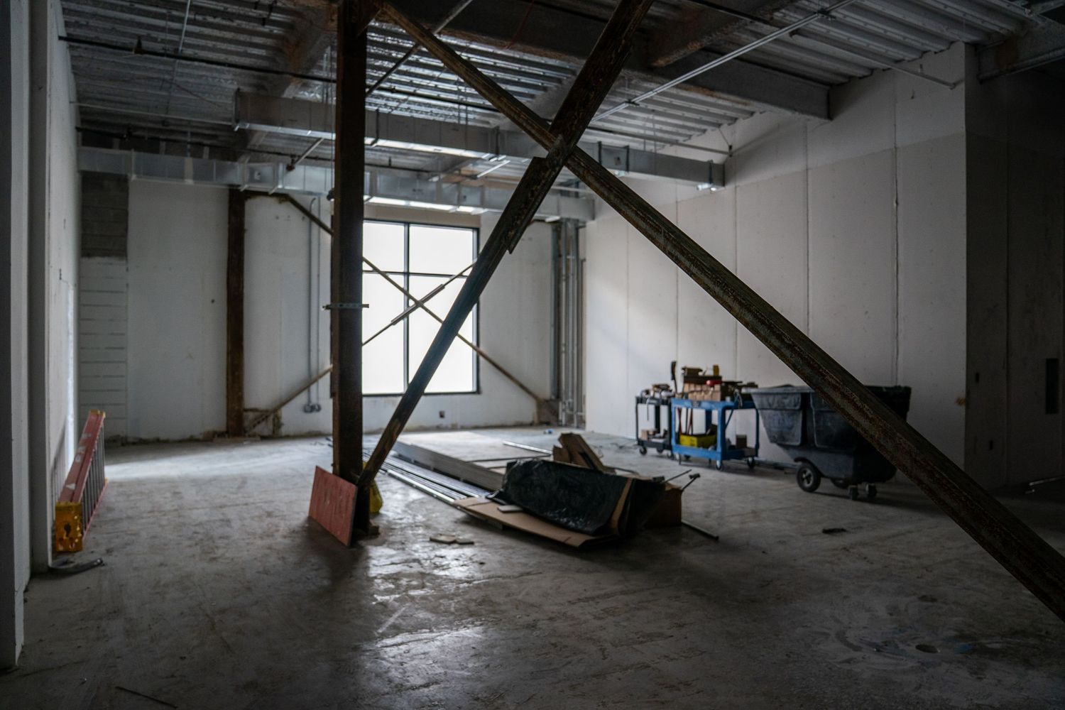 Interior view of an unfinished commercial space with exposed beams and construction materials scattered on concrete floor.