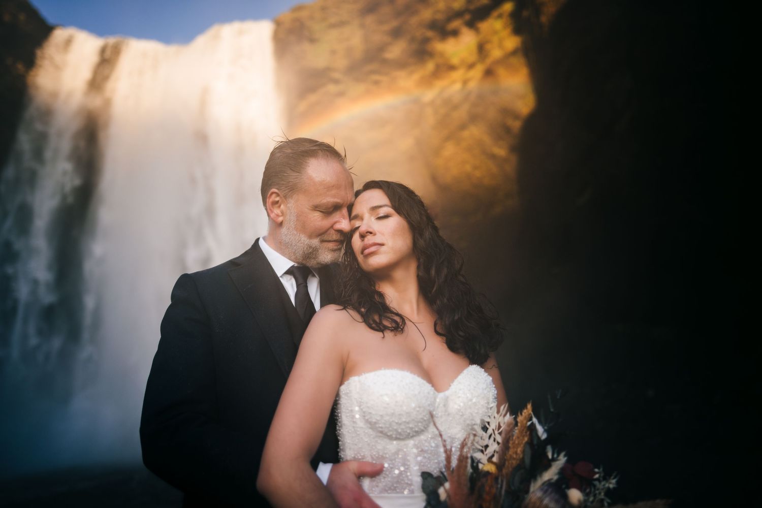 Iceland destination wedding romantic couple embraces in front of a majestic waterfall at sunset with golden light illuminating the scene.
