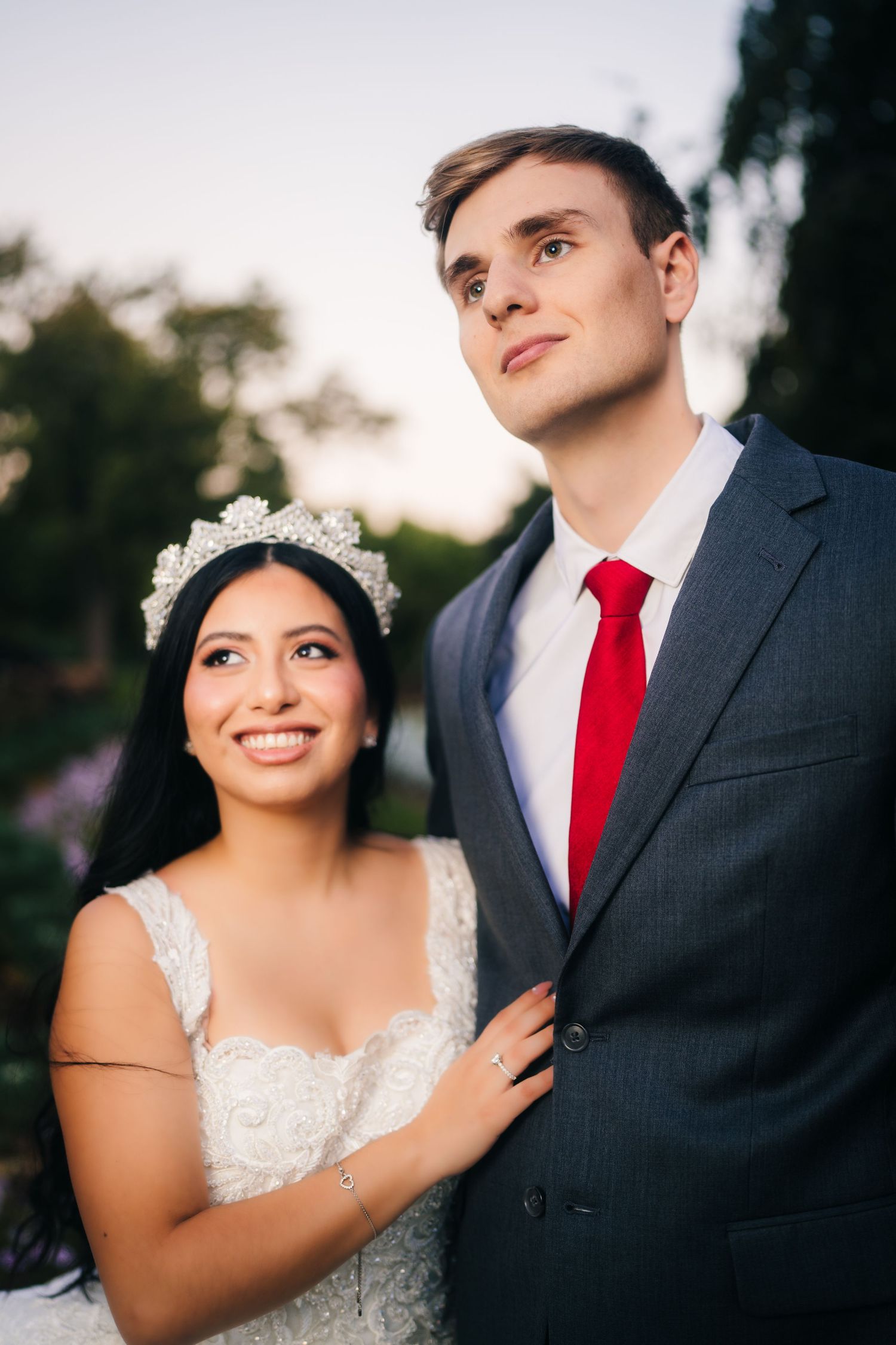 A bride in white lace dress and groom in dark suit with red tie pose together for romantic wedding portraits outdoors.