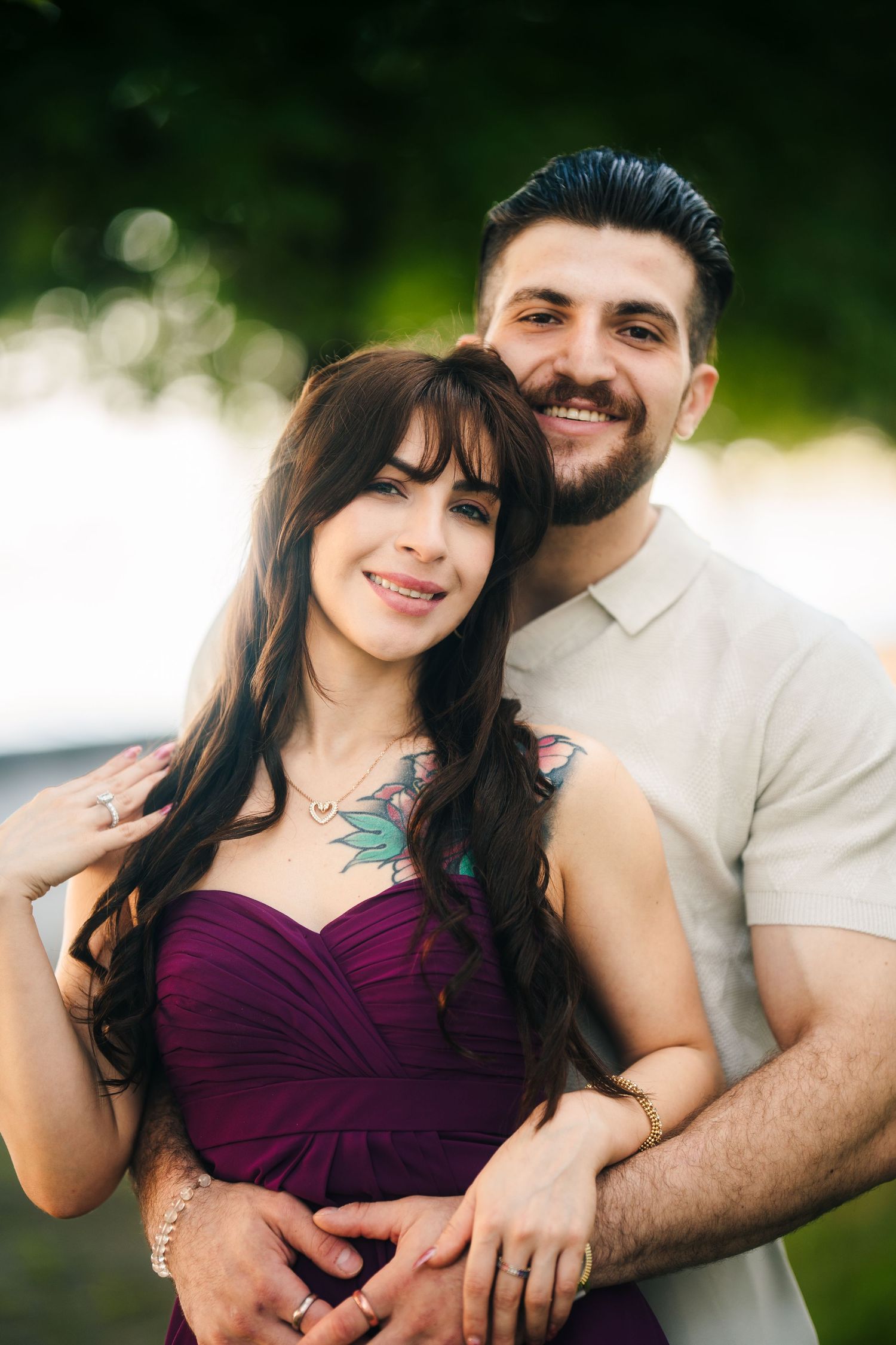 A couple embraces outdoors in romantic portrait, with her in a purple dress and him in a beige shirt.