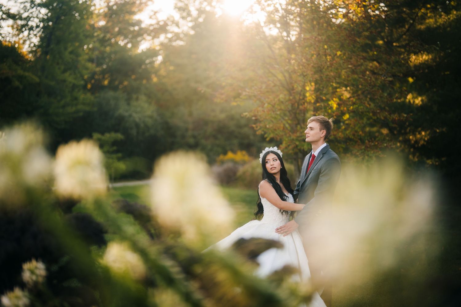 Oakville park wedding. A romantic wedding couple embraces in a sun-drenched garden with soft flowers in the foreground during golden hour.