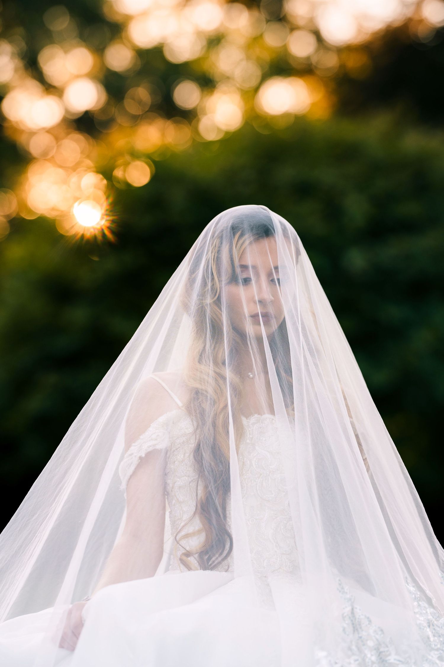 A bride in a white veil stands in front of bokeh lights during golden hour sunset.