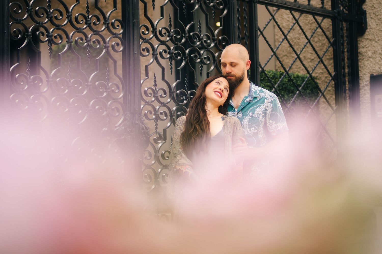 A couple poses together in front of an ornate wrought iron gate with blurred pink flowers in the background.