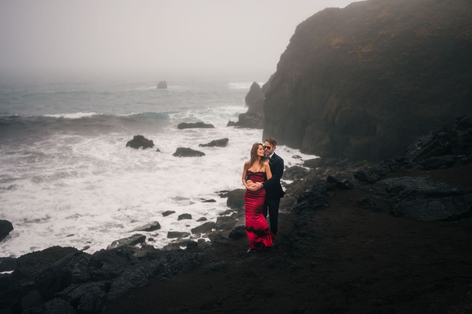 A couple embraces on dramatic rocky coastline with crashing waves and misty cliffs in the background.