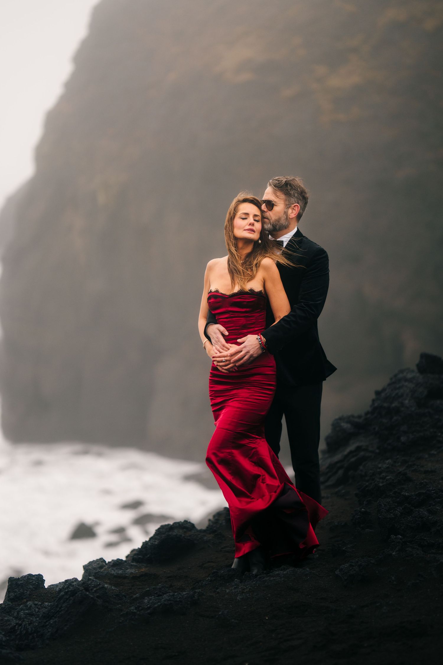 Iceland destination wedding. A couple embraces on dramatic black rocks against misty coastal cliffs in four romantic engagement photos.