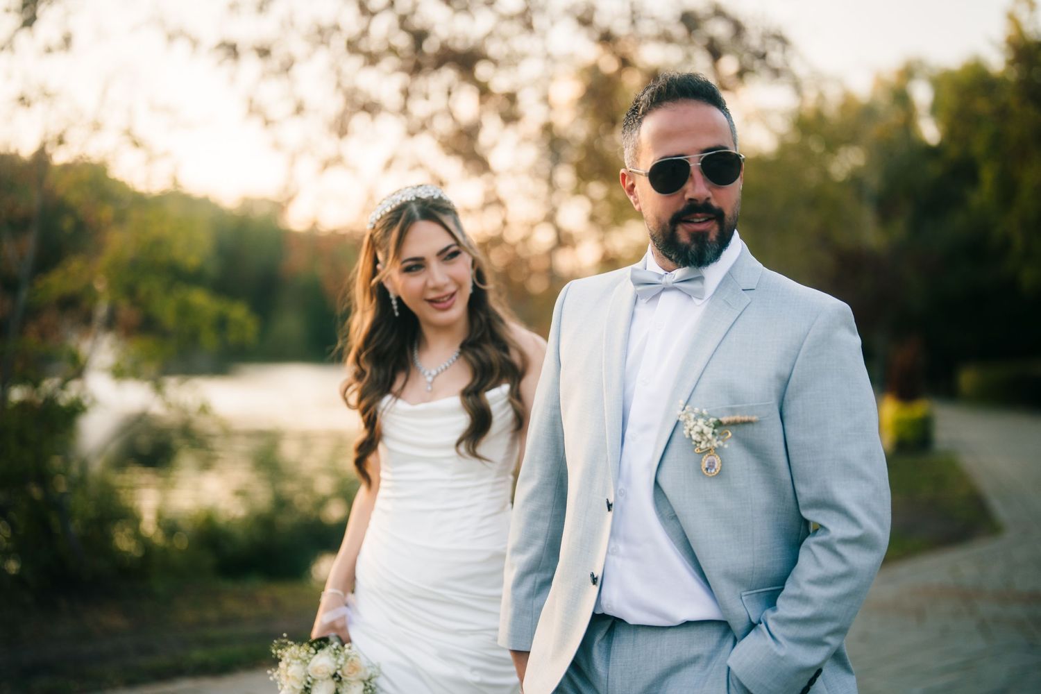 A bride in white gown and groom in light blue suit pose together outdoors at sunset.