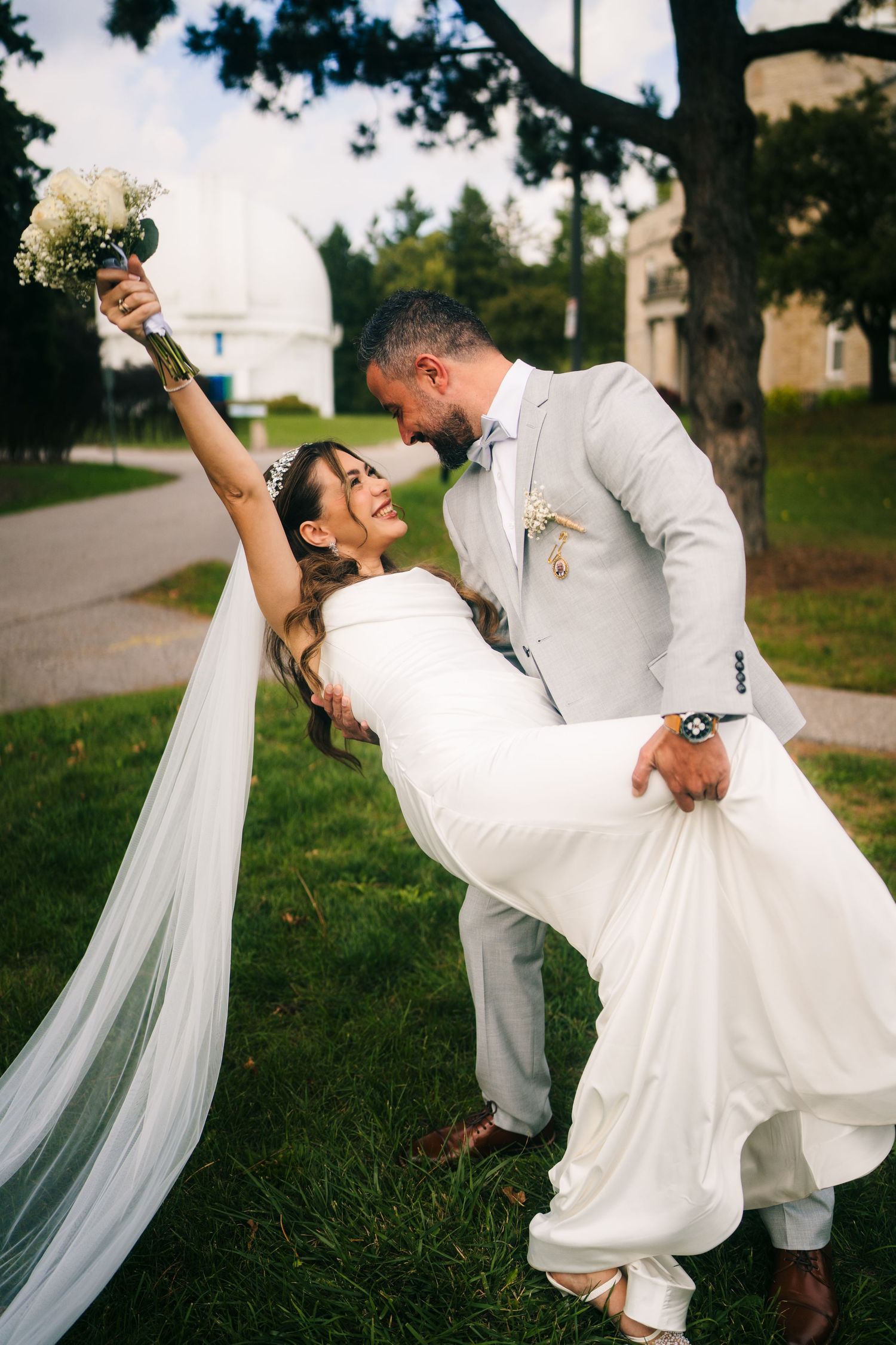 A romantic wedding couple performs a dramatic dip pose on a grassy lawn, with the bride's veil flowing elegantly behind them.