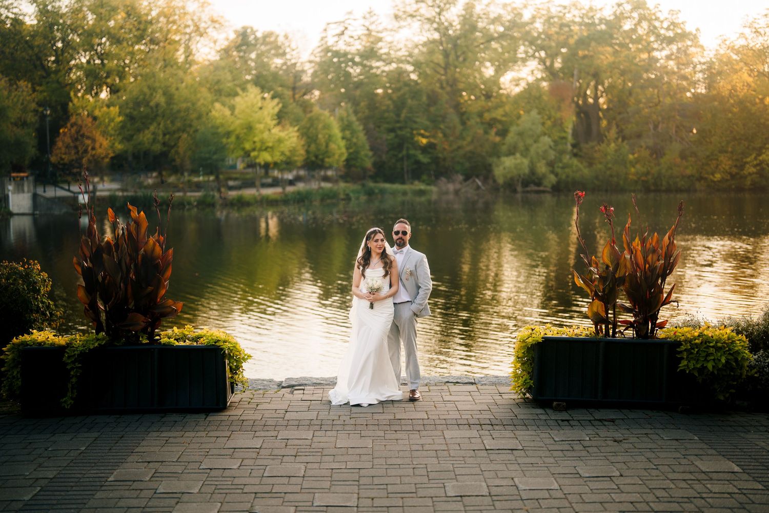 A couple walks along a scenic lakeside path at sunset while surrounded by lush autumn foliage and golden light.
