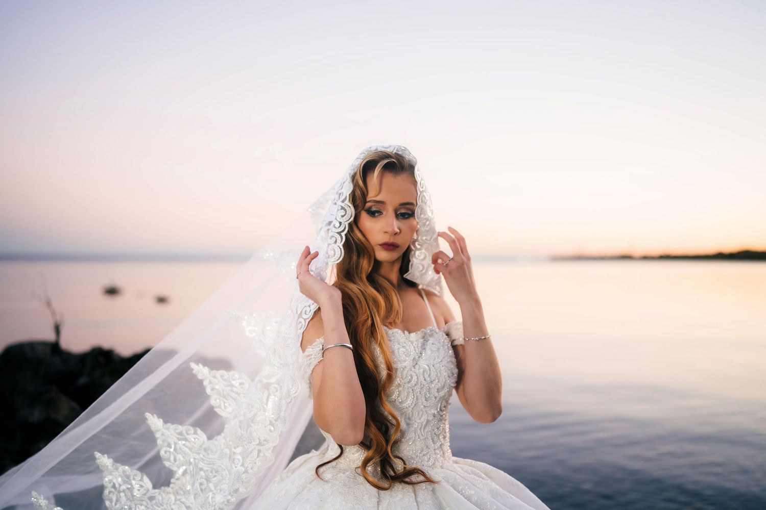 Brueckner Rhododendron Gardens bride in a white lace gown poses by a serene lake at sunset with her veil flowing in the breeze.