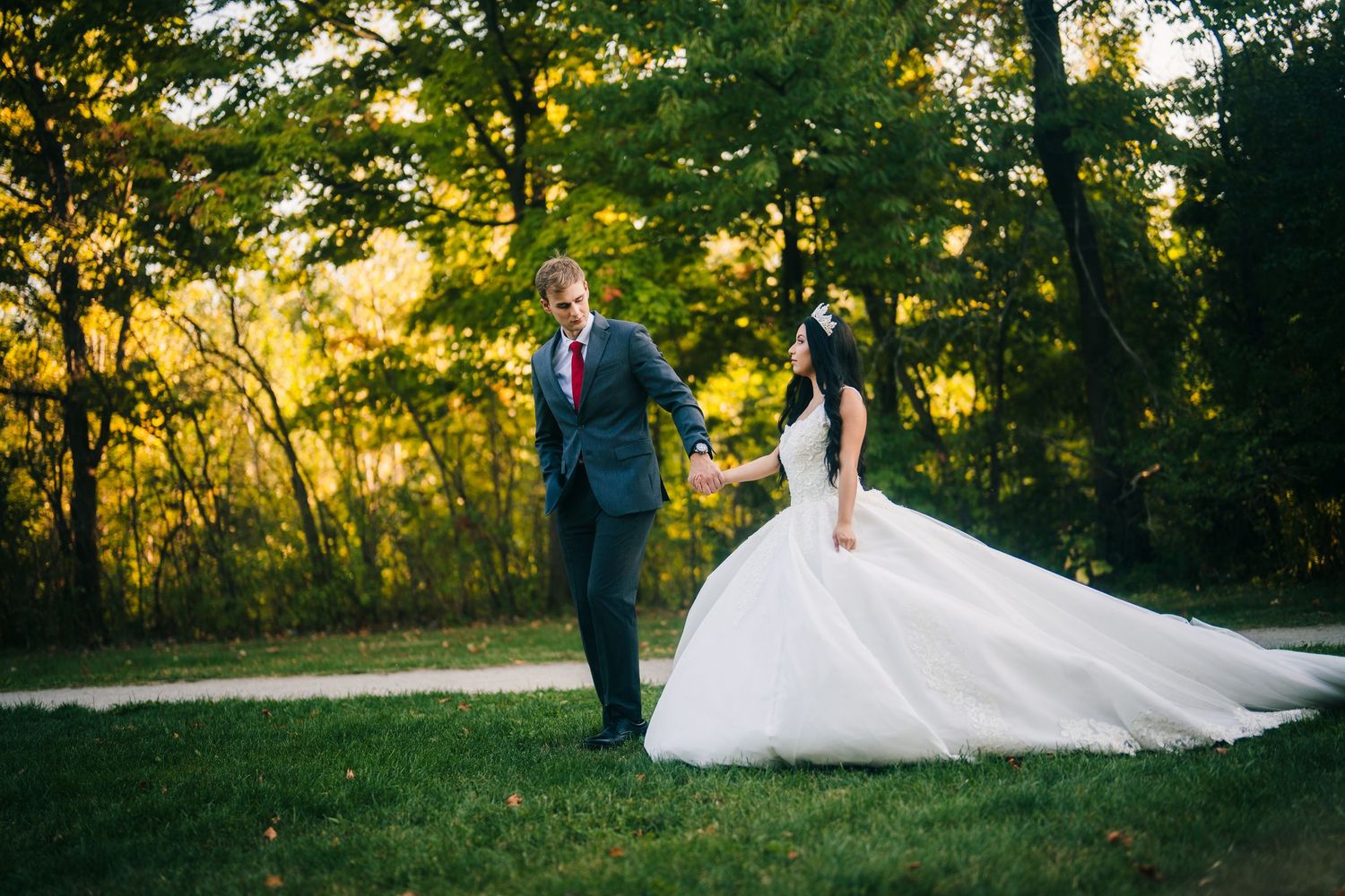 Oakville Newlywed couple walks through a sunlit forest during golden hour as bride's white dress trails behind them.