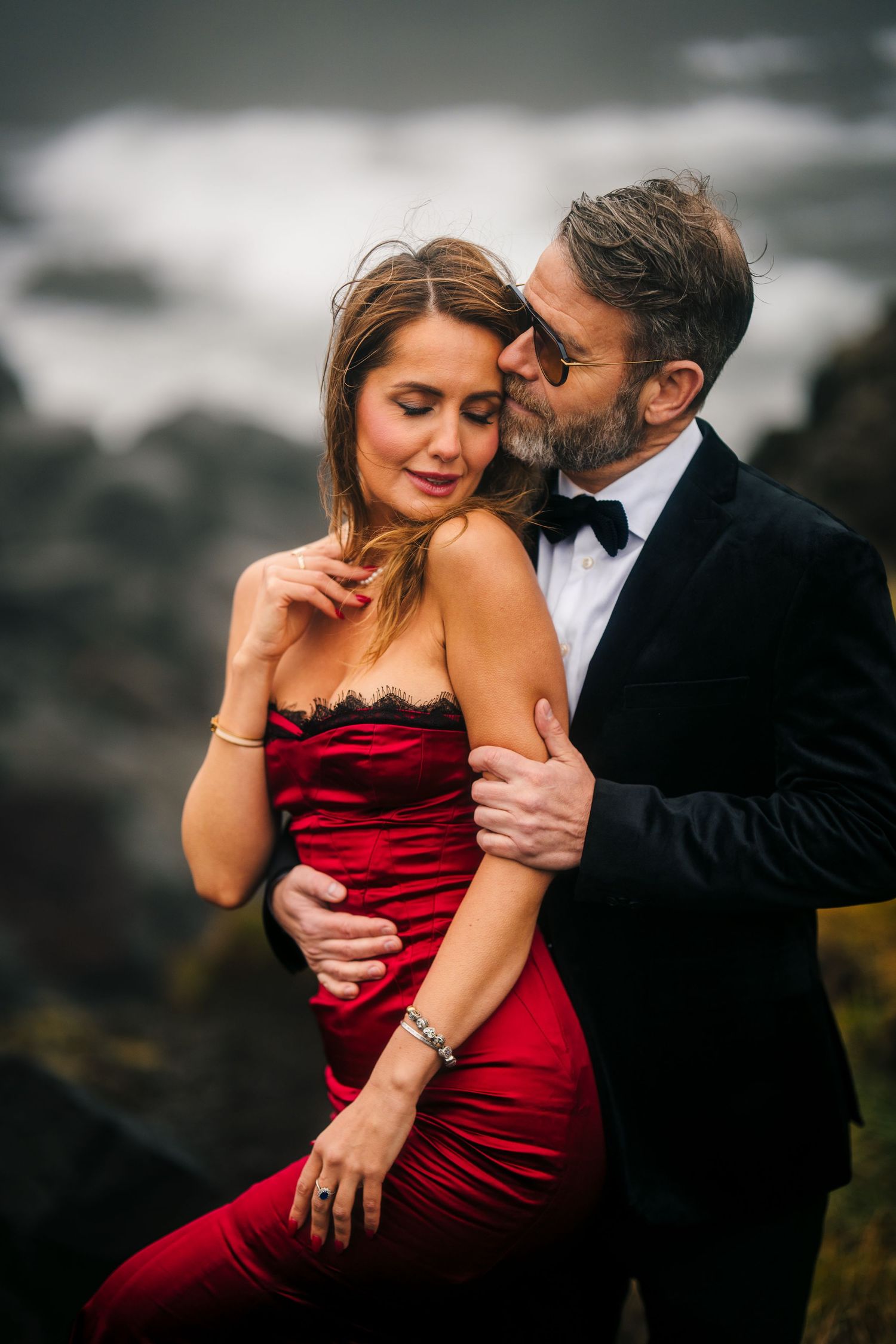 Iceland destination wedding. A romantic couple in formal attire embrace against a moody backdrop, with the woman wearing a red velvet dress.