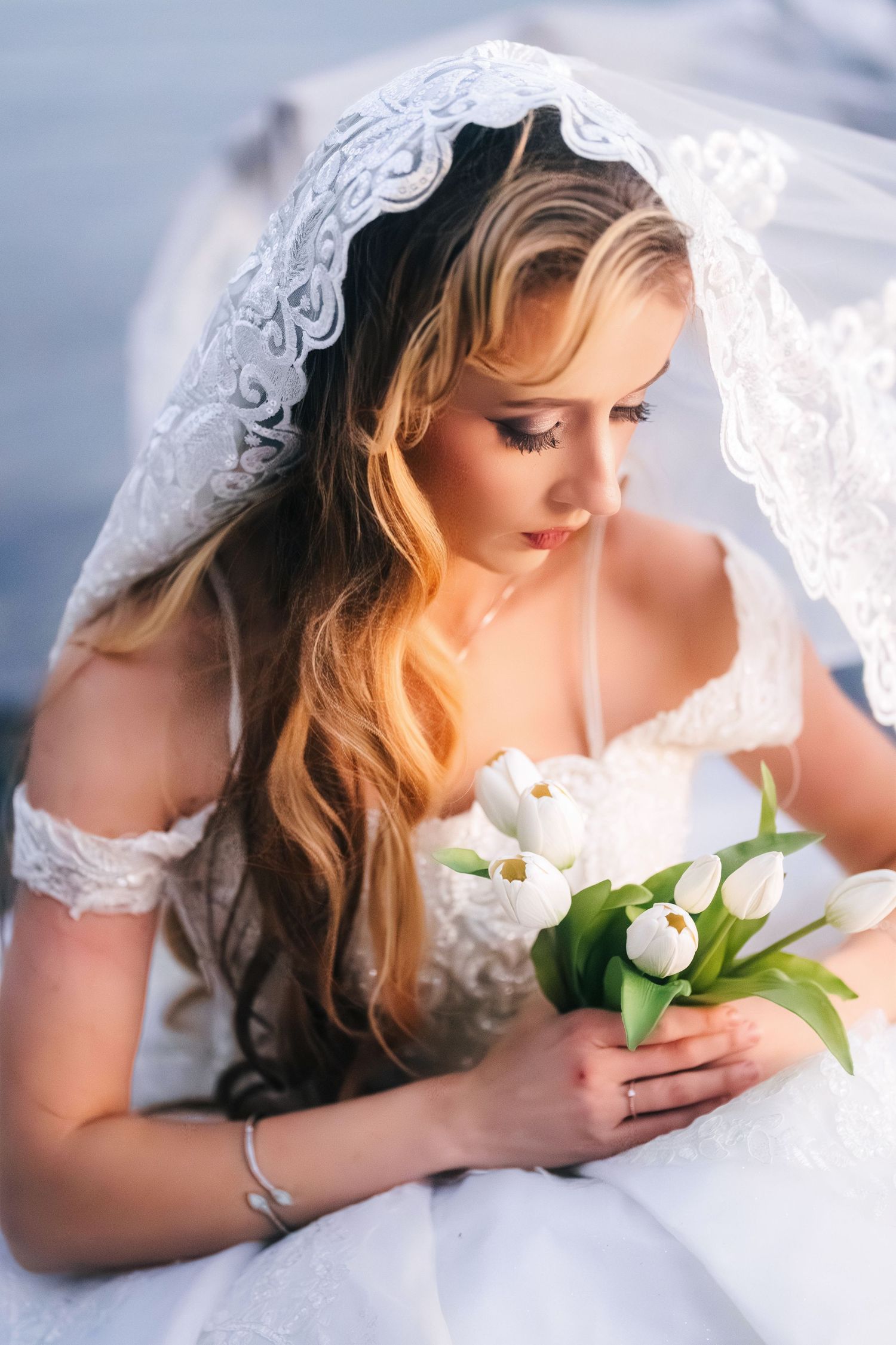 Bride in delicate lace veil holds white tulip bouquet while gazing down in soft natural lighting.