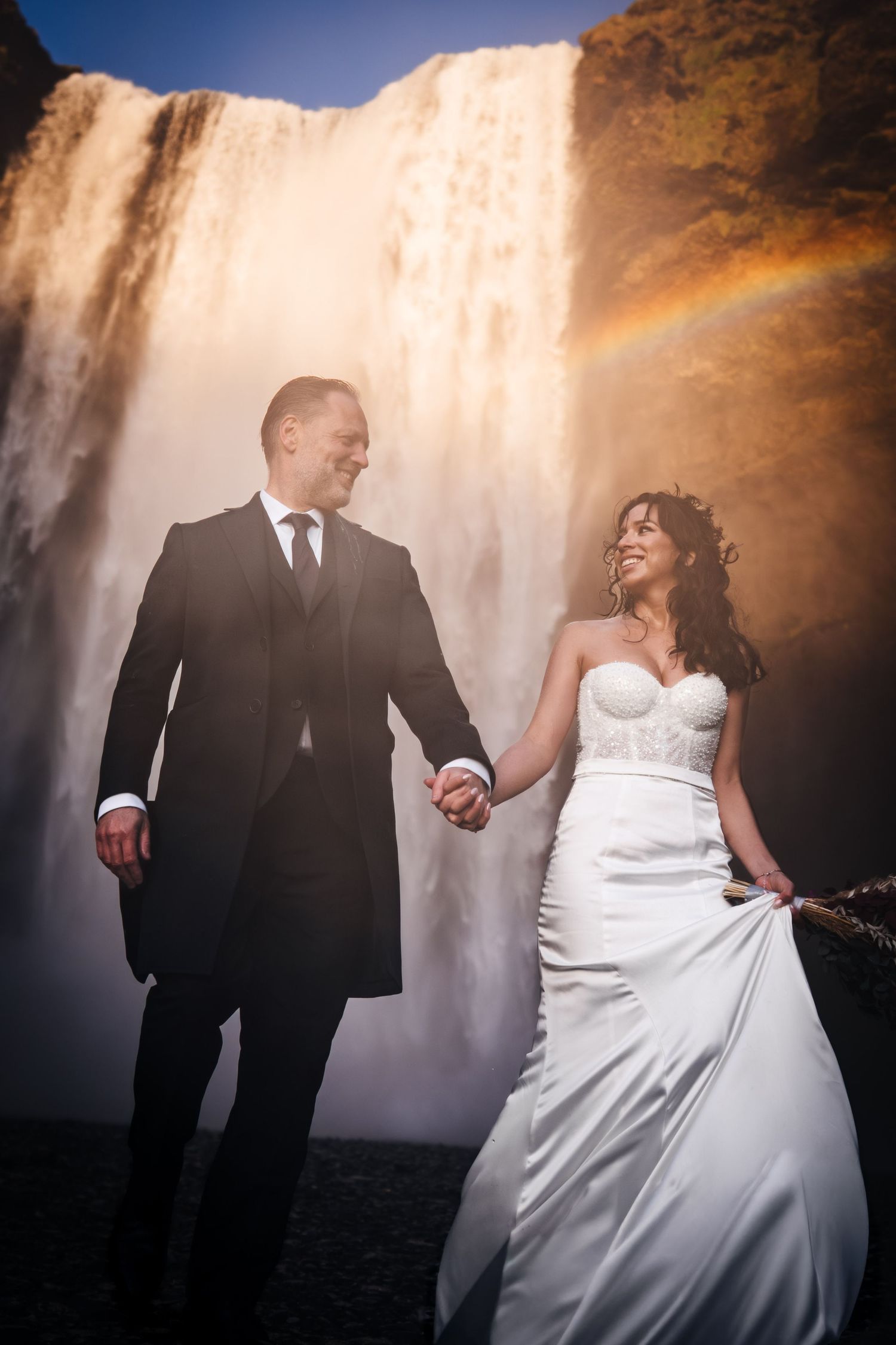 Iceland destination wedding. A romantic wedding couple poses against a waterfall backdrop as sunlight creates a golden glow in the mist.
