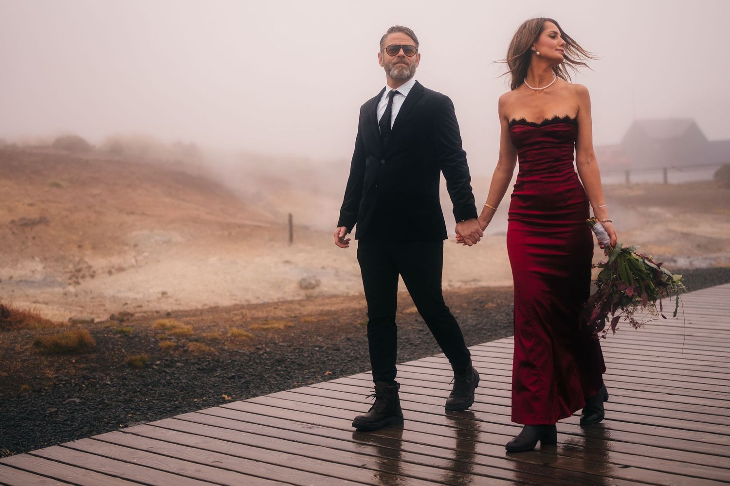 Iceland destination wedding. A couple in formal attire walks along a misty boardwalk, with the woman wearing a burgundy dress.