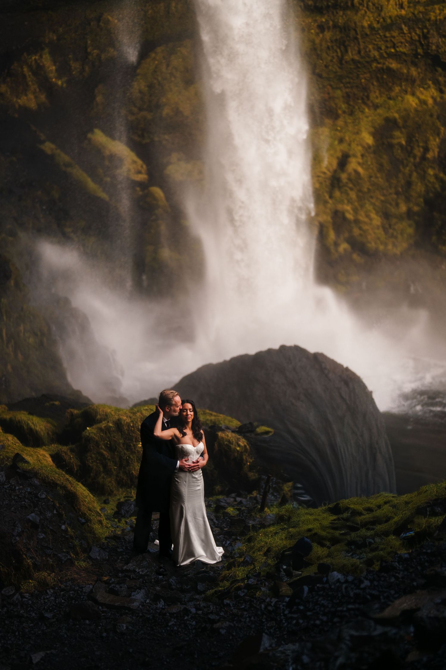 Iceland destination wedding. A couple embraces against the dramatic backdrop of a misty waterfall and mossy rocks at sunset.