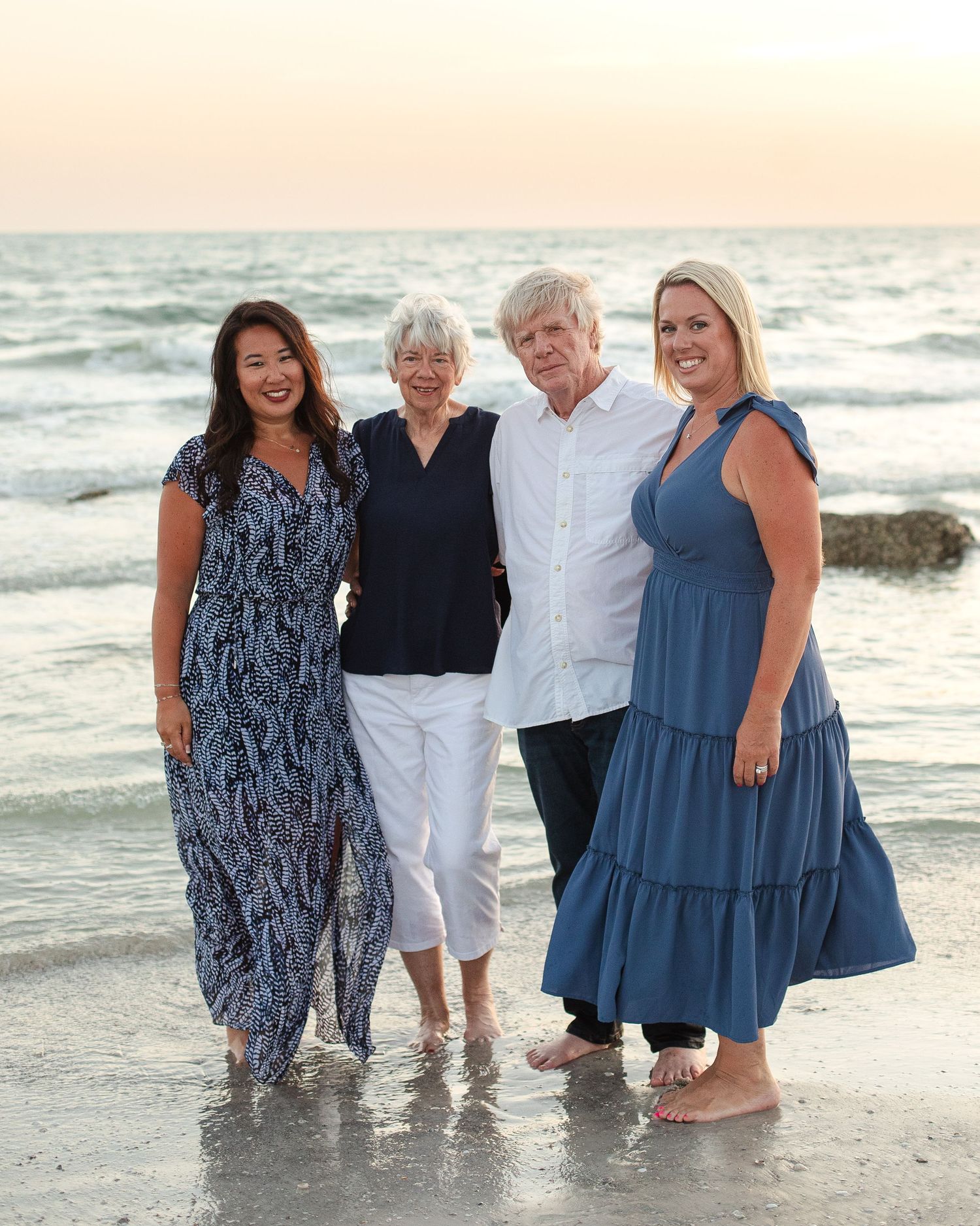 Four women smiling together on a beach at sunset, wearing blue and white outfits.