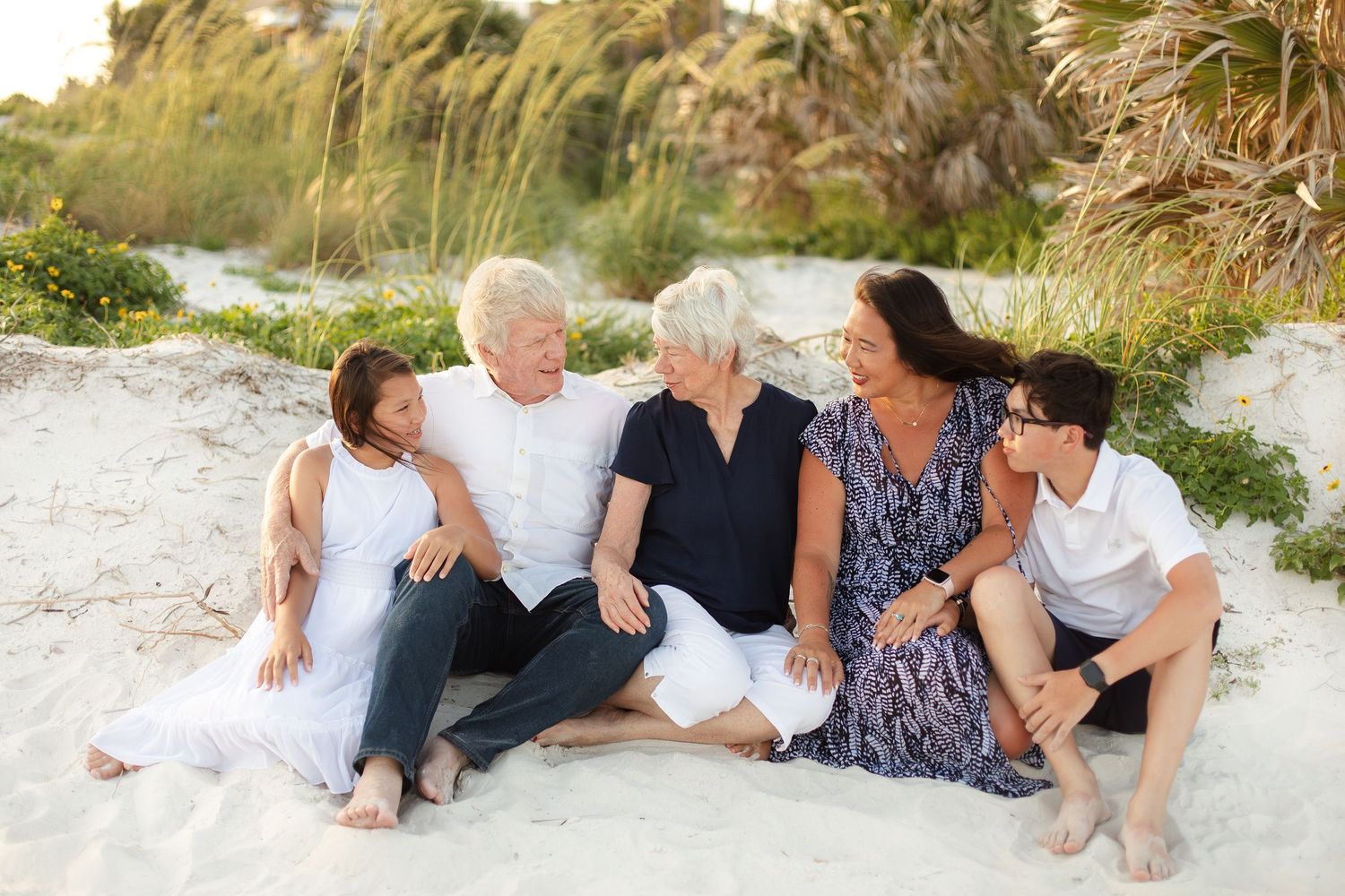 Family of five smiling together on a sandy beach, surrounded by lush greenery on a sunny day.