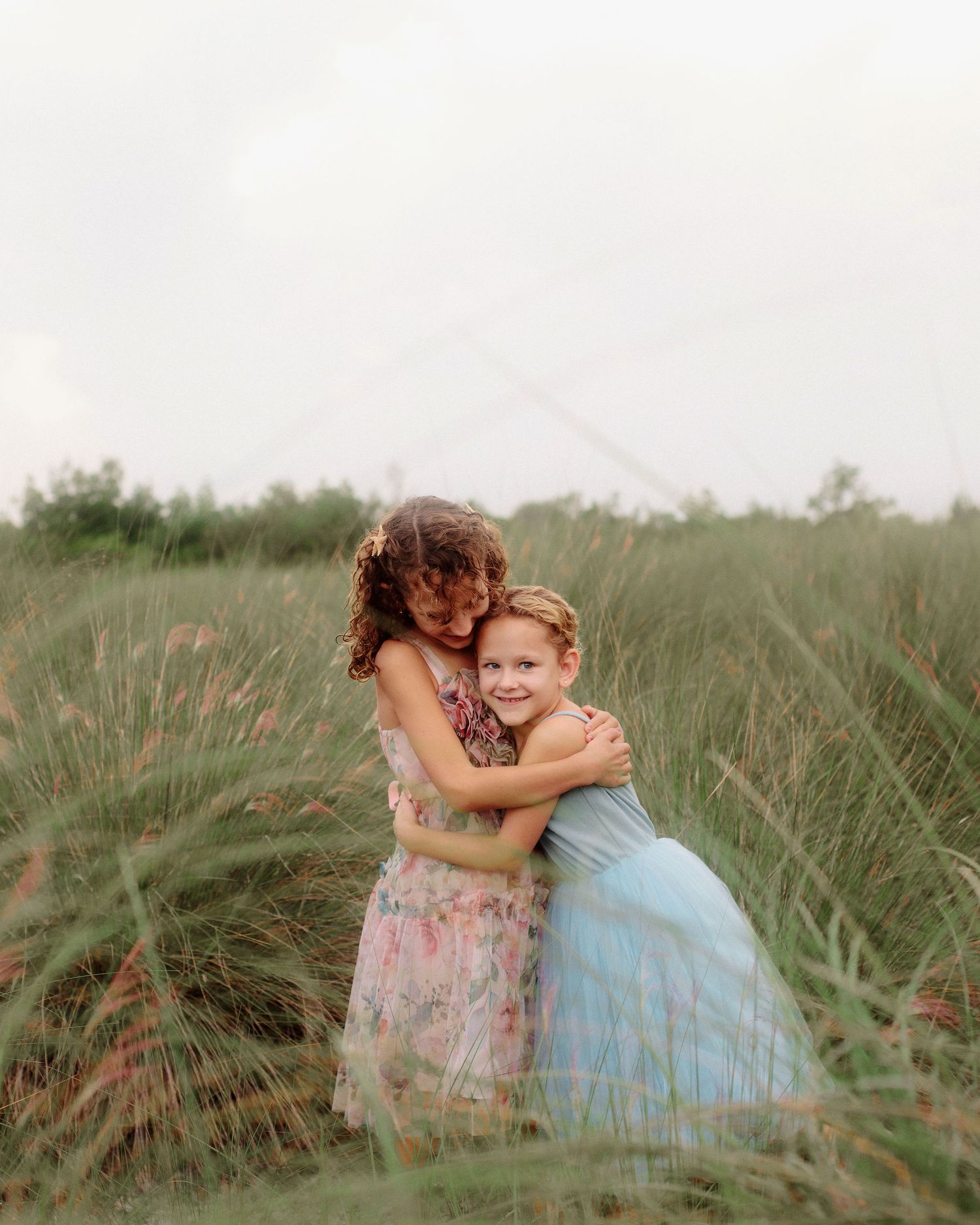 Two young girls in pastel floral and blue tulle dresses hug tightly in a tall grassy field under a soft, overcast sky.