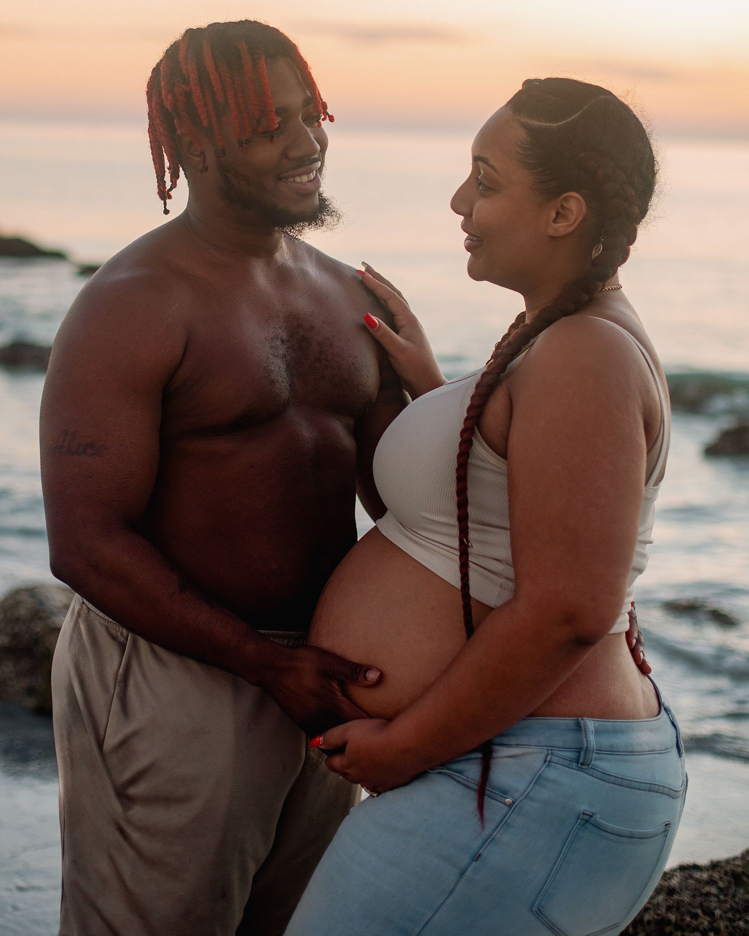 Pregnant couple embracing on rocky beach at sunset, woman in white crop top and jeans, man shirtless in gray pants.