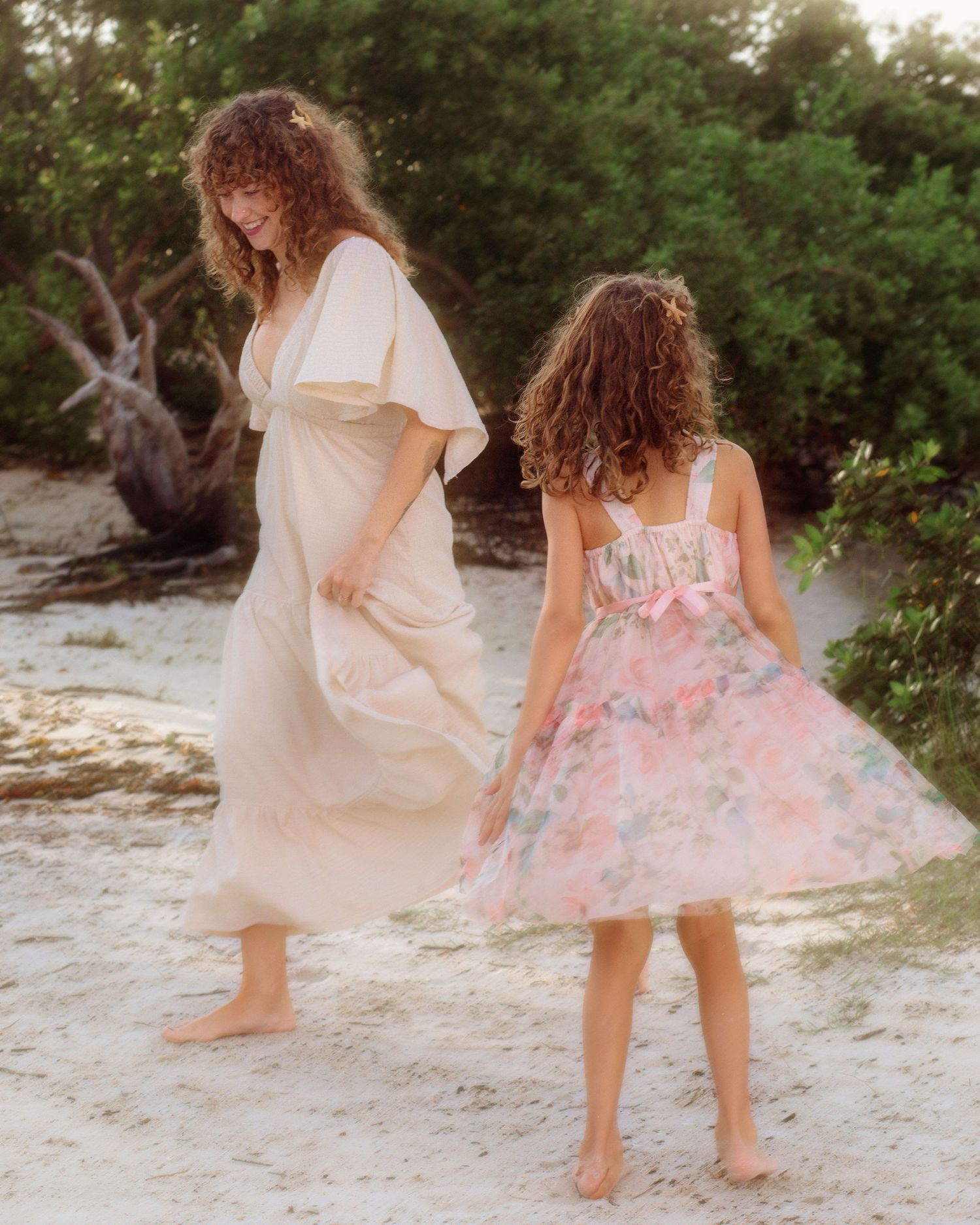 Woman in a cream flowing dress and girl in a pink floral tulle dress walk barefoot on a sandy beach near green trees.