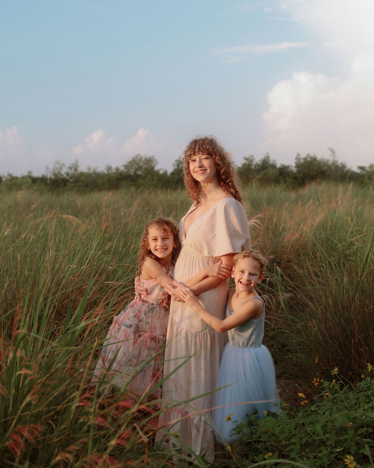 A woman in a cream dress stands with two young girls in a tall grass field at golden hour, embracing as soft clouds fill the sky.