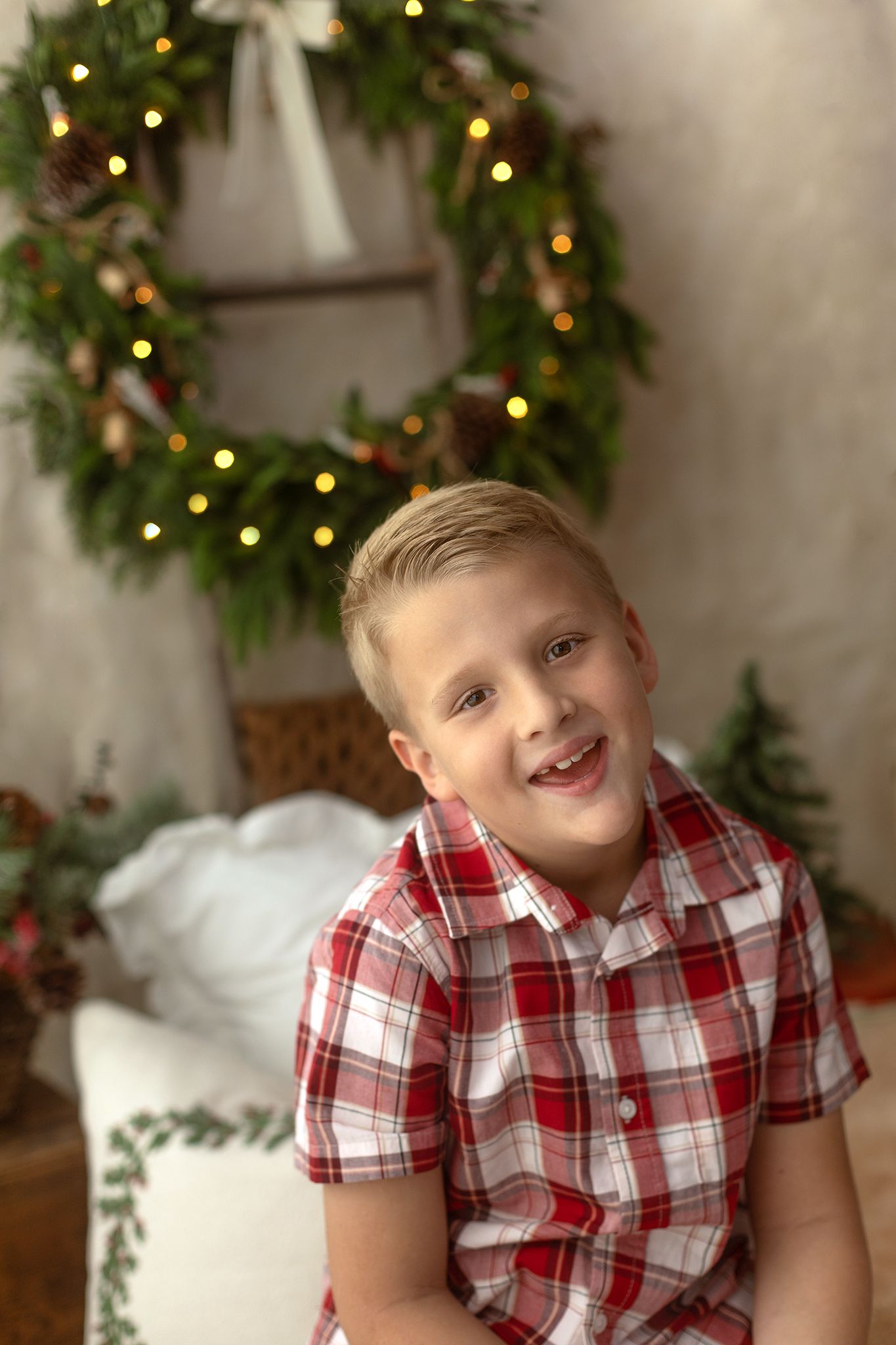 Smiling boy in red plaid shirt poses in front of festive Christmas wreath with warm glowing lights.