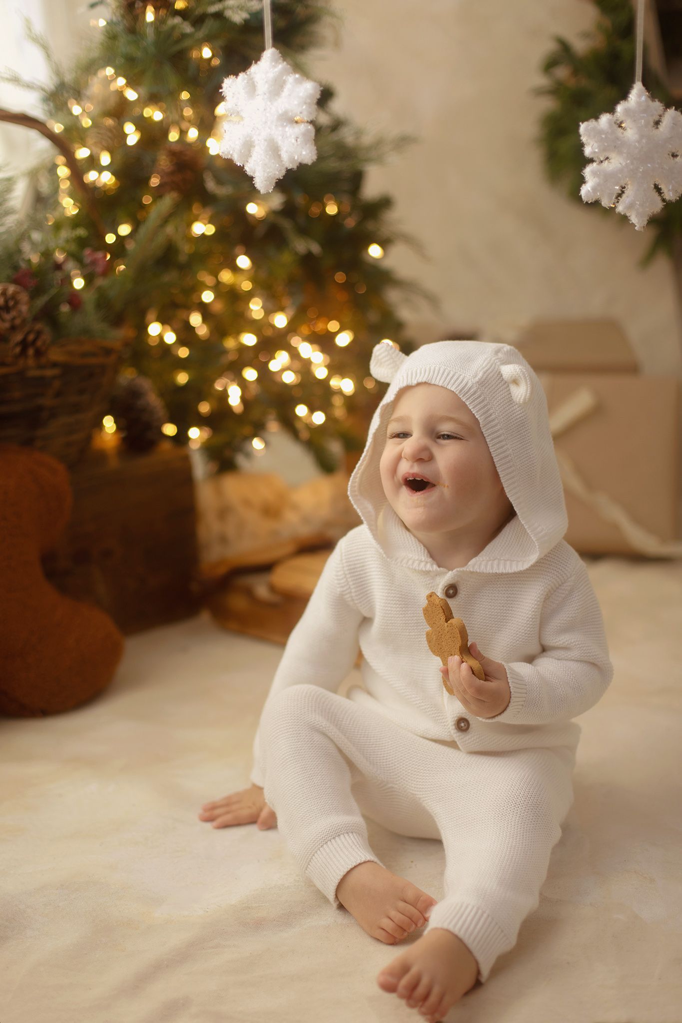 Happy baby in white bear knit onesie sits on fur rug holding gingerbread cookie near glowing Christmas tree.