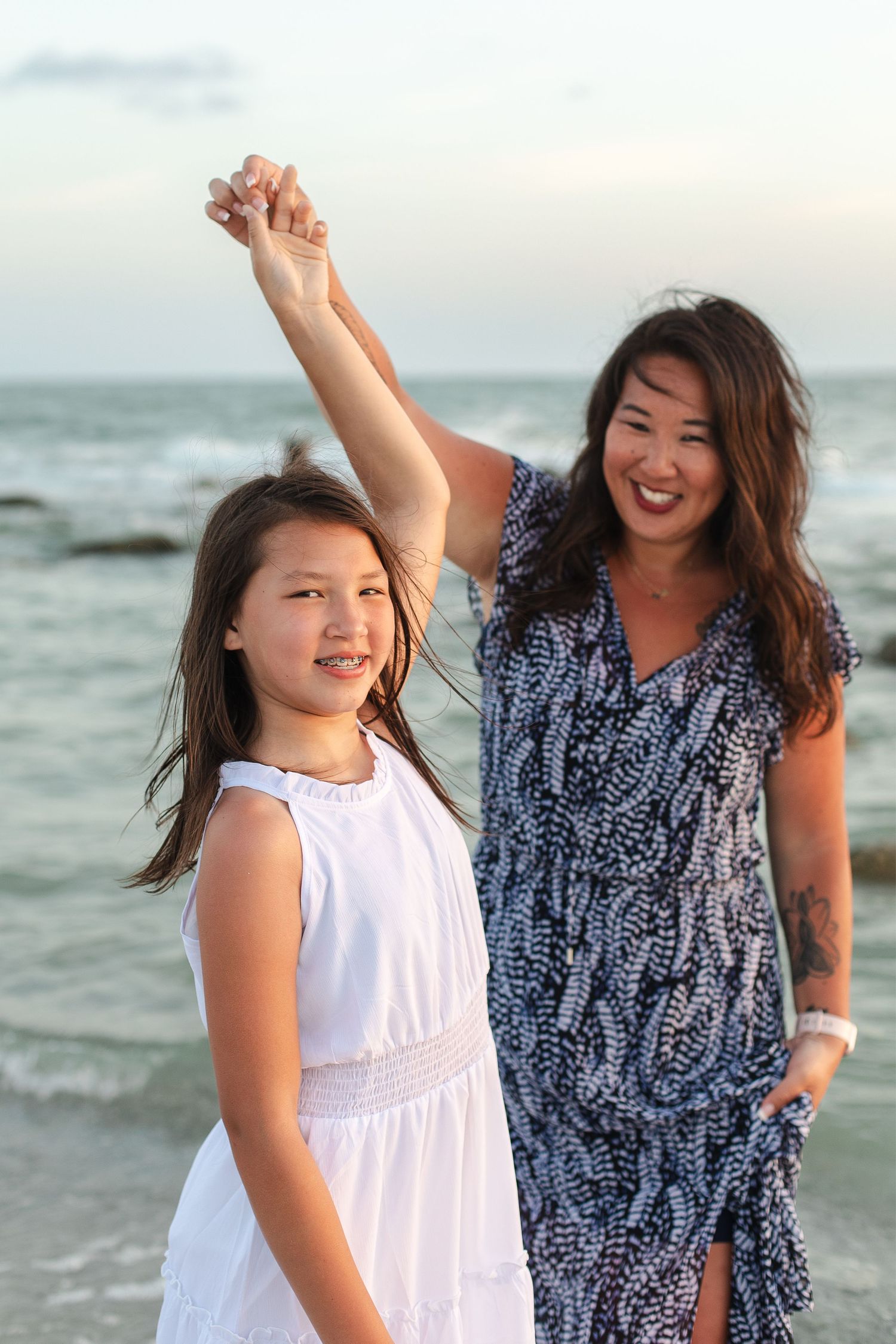 Mother and daughter holding hands on beach, smiling in white and blue dresses at sunset.