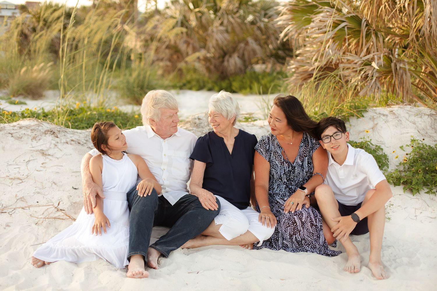 Multi-generational family of five smiling together on a sandy beach surrounded by palm trees and dunes.