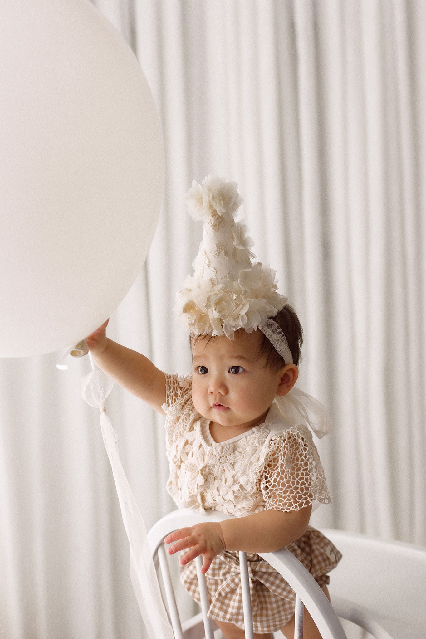 Baby in a cream floral party hat and lace outfit holding a large white balloon while standing on a white chair against soft curtains.