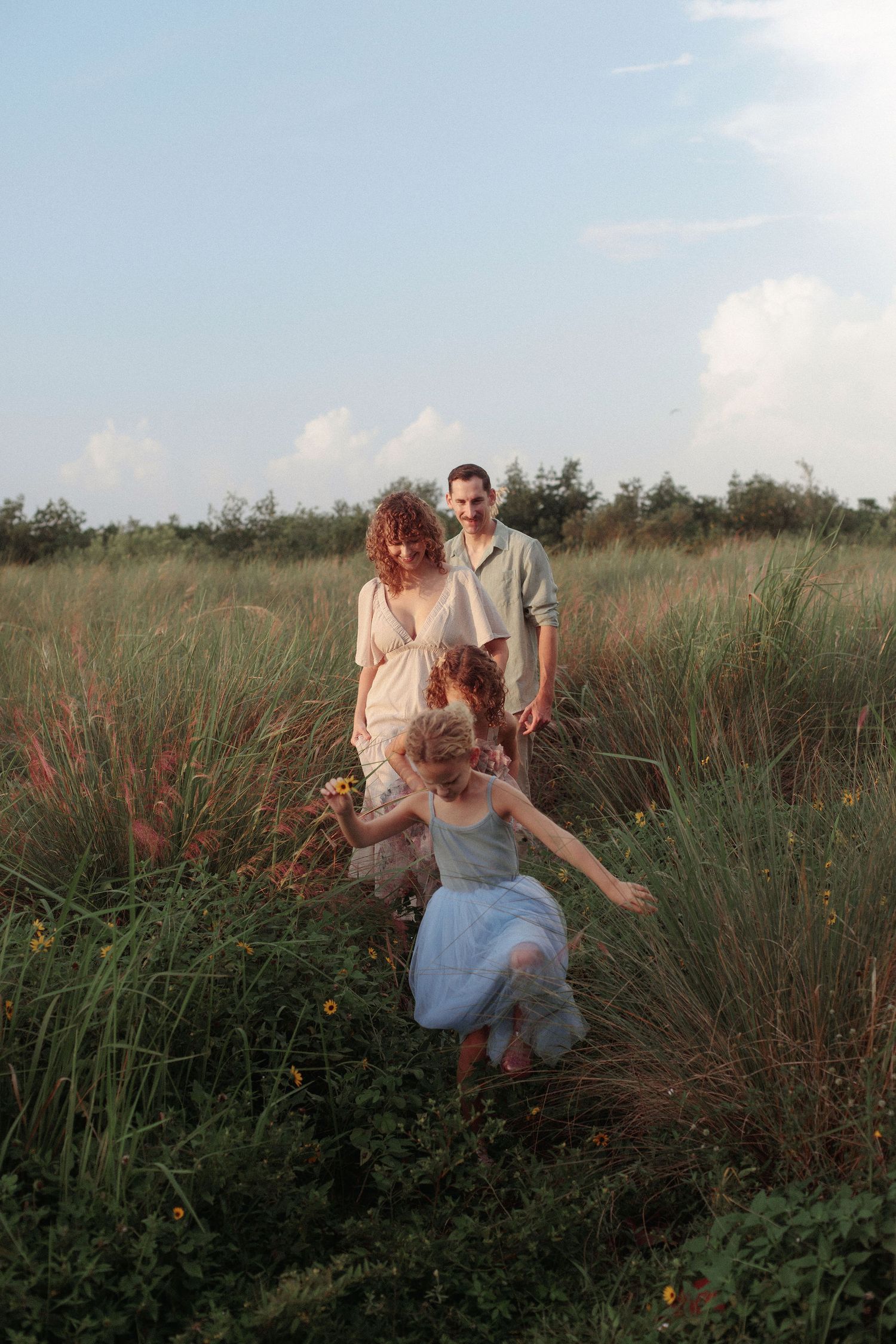 A family walks through tall golden grass and wildflowers at sunset as a young girl in a blue dress runs ahead of her parents.