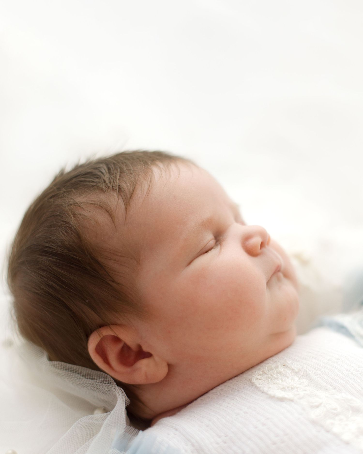 Close-up profile of sleeping newborn baby with dark hair wrapped in soft white fabric.