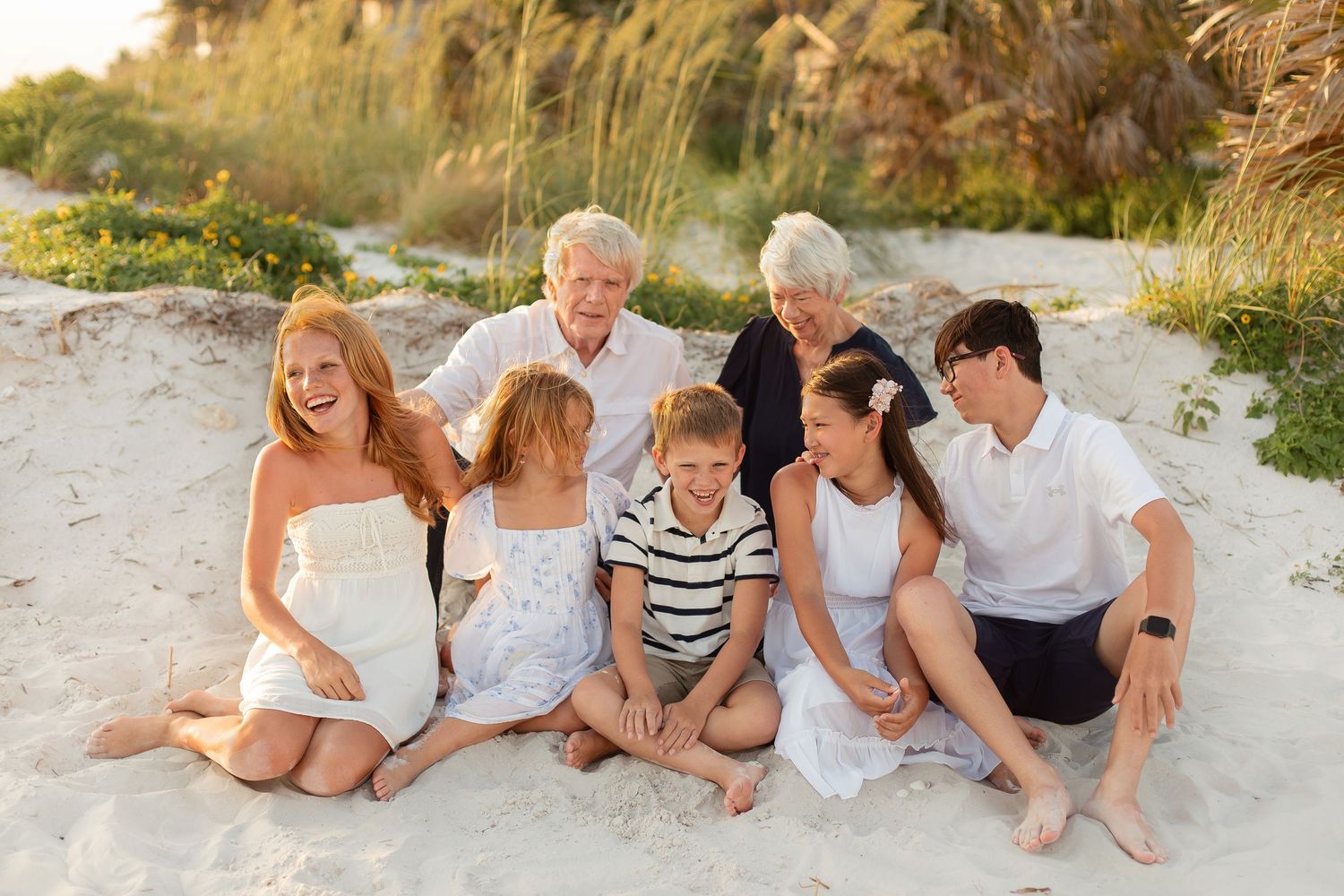 Multi-generational family laughing together on sandy beach at golden hour, dressed in white and navy outfits.