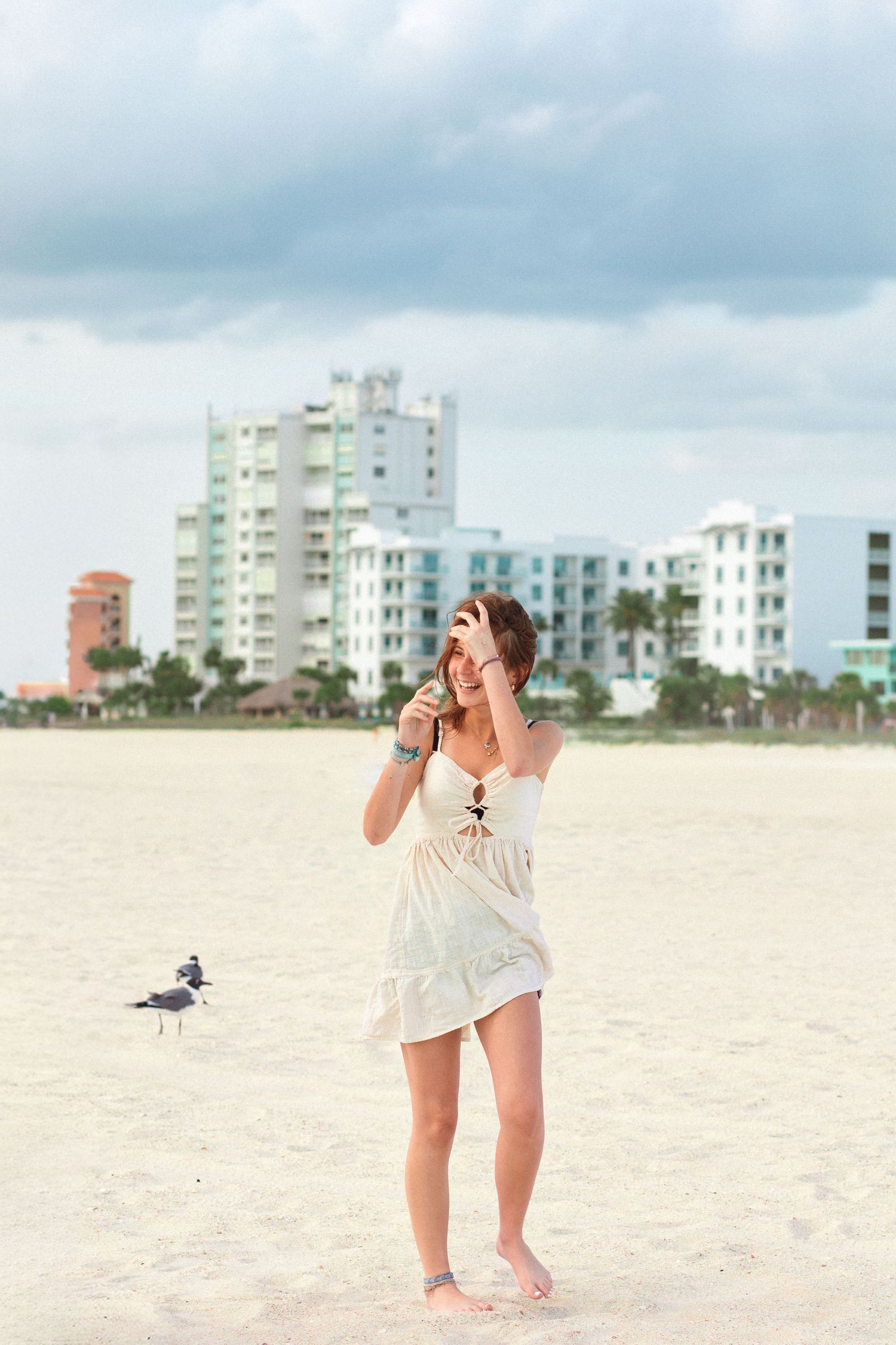 Young woman in white sundress standing barefoot on sandy beach with seagull and coastal buildings in background.