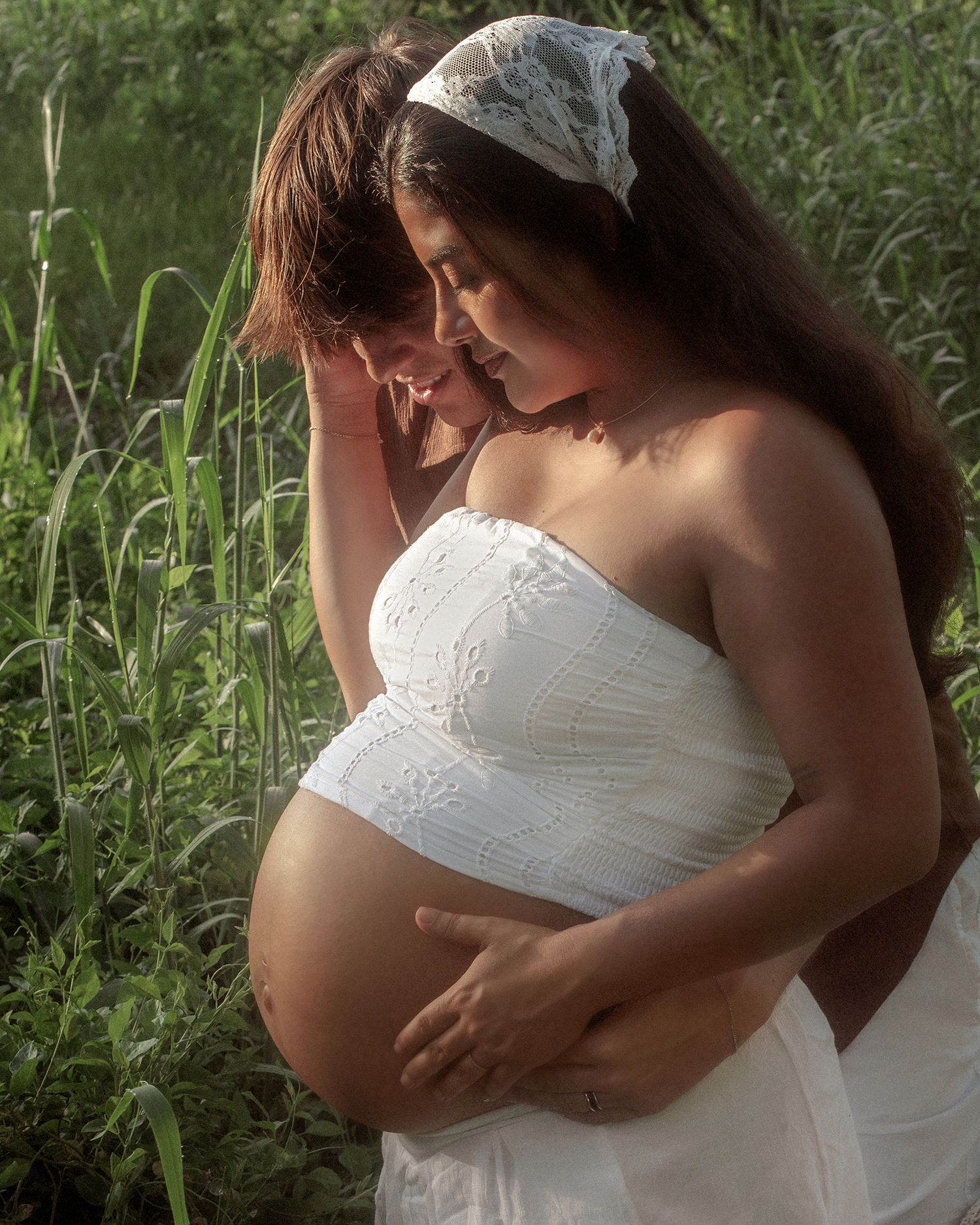 Pregnant woman in white strapless dress embraced by partner outdoors among lush green foliage.