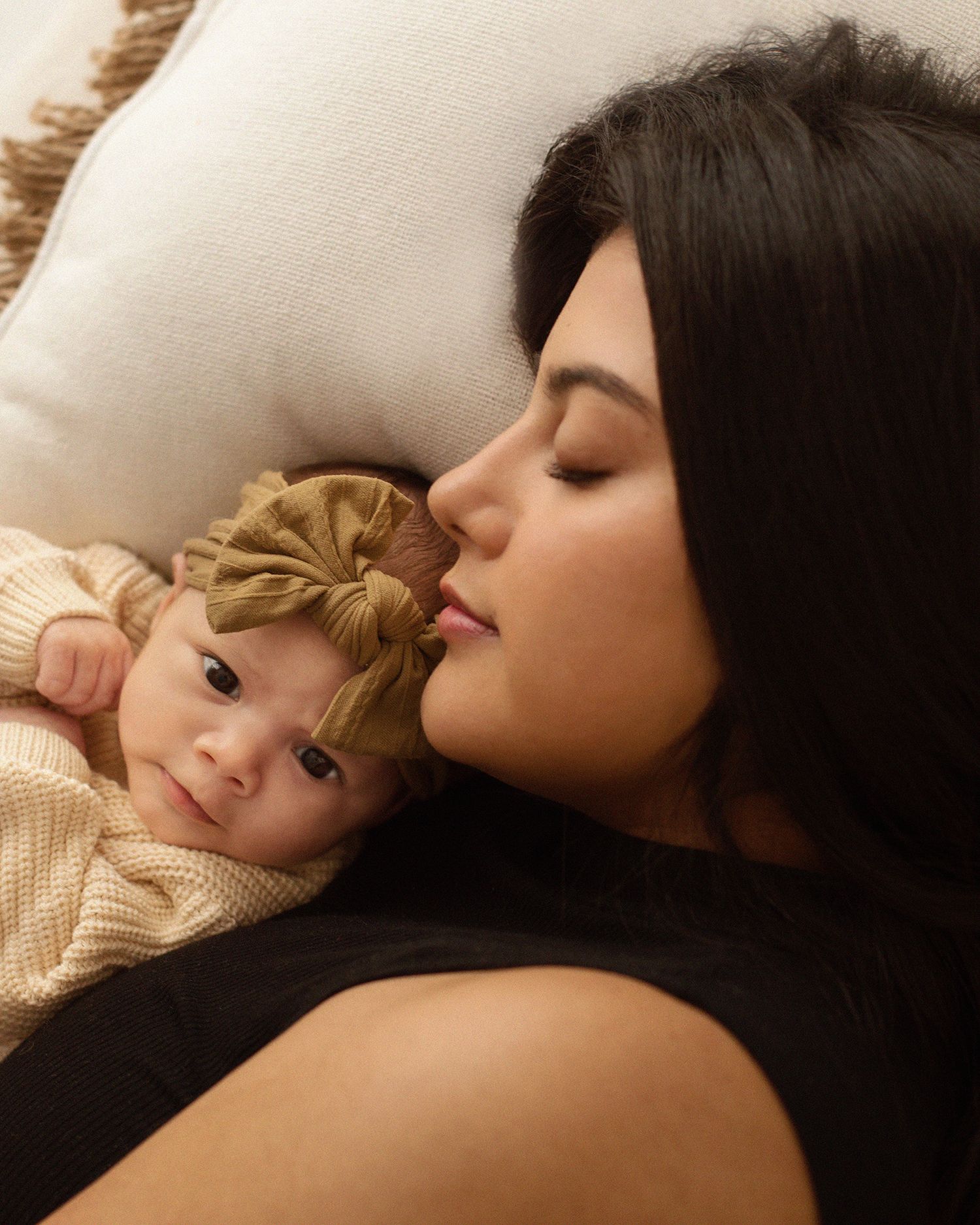 A dark-haired woman lovingly nuzzles her newborn baby girl wearing a golden bow headband on a white pillow.