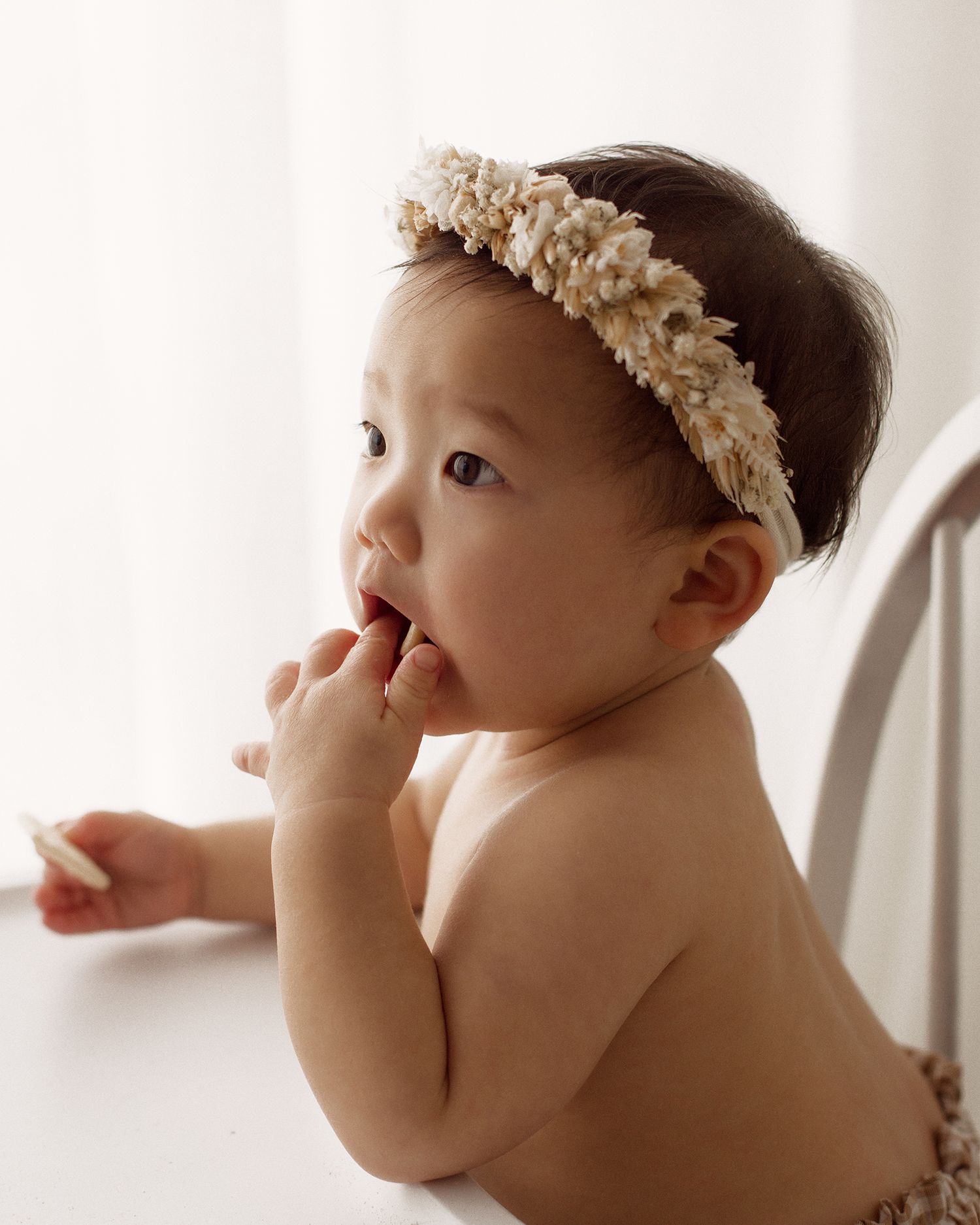 Baby wearing a cream floral headband chews on their fingers while leaning on a white high chair tray in soft natural light.