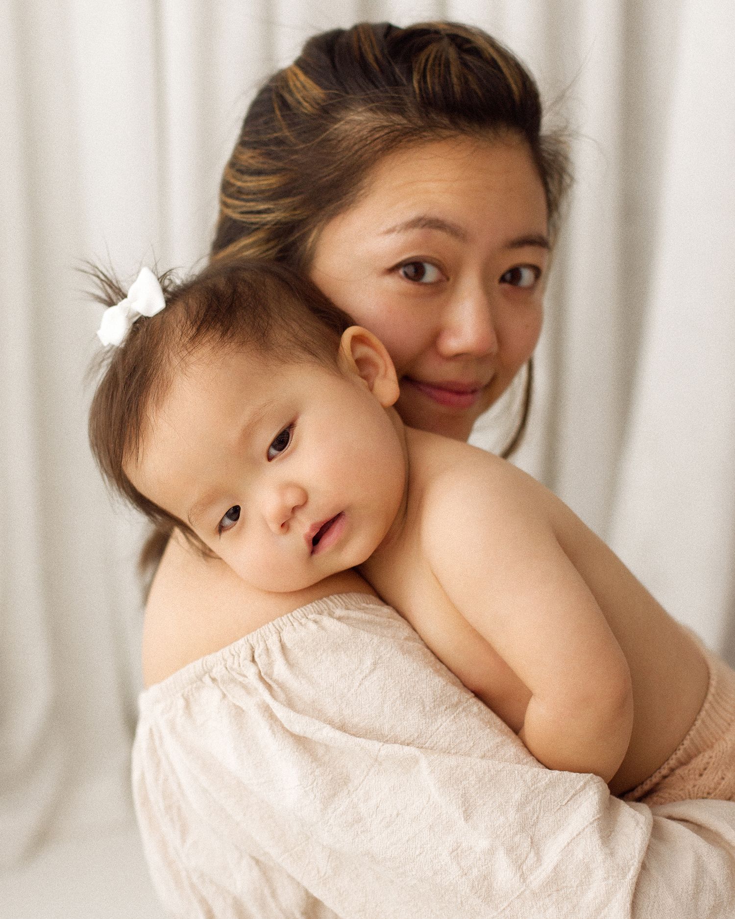 A smiling woman with dark hair holds a baby wearing a white bow against her shoulder in a soft, light-filled studio portrait.