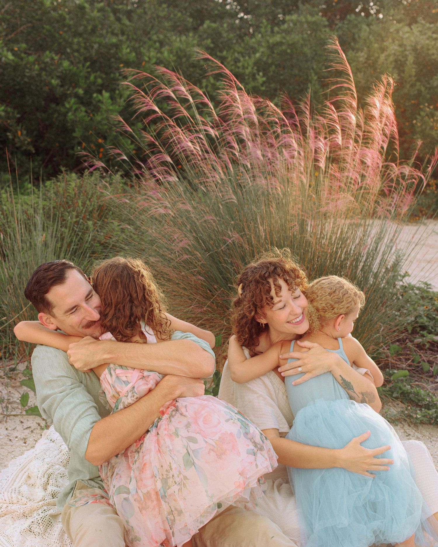 A smiling family of four in soft pastel outfits hugs on a blanket outdoors, surrounded by green foliage and pink muhly grass.